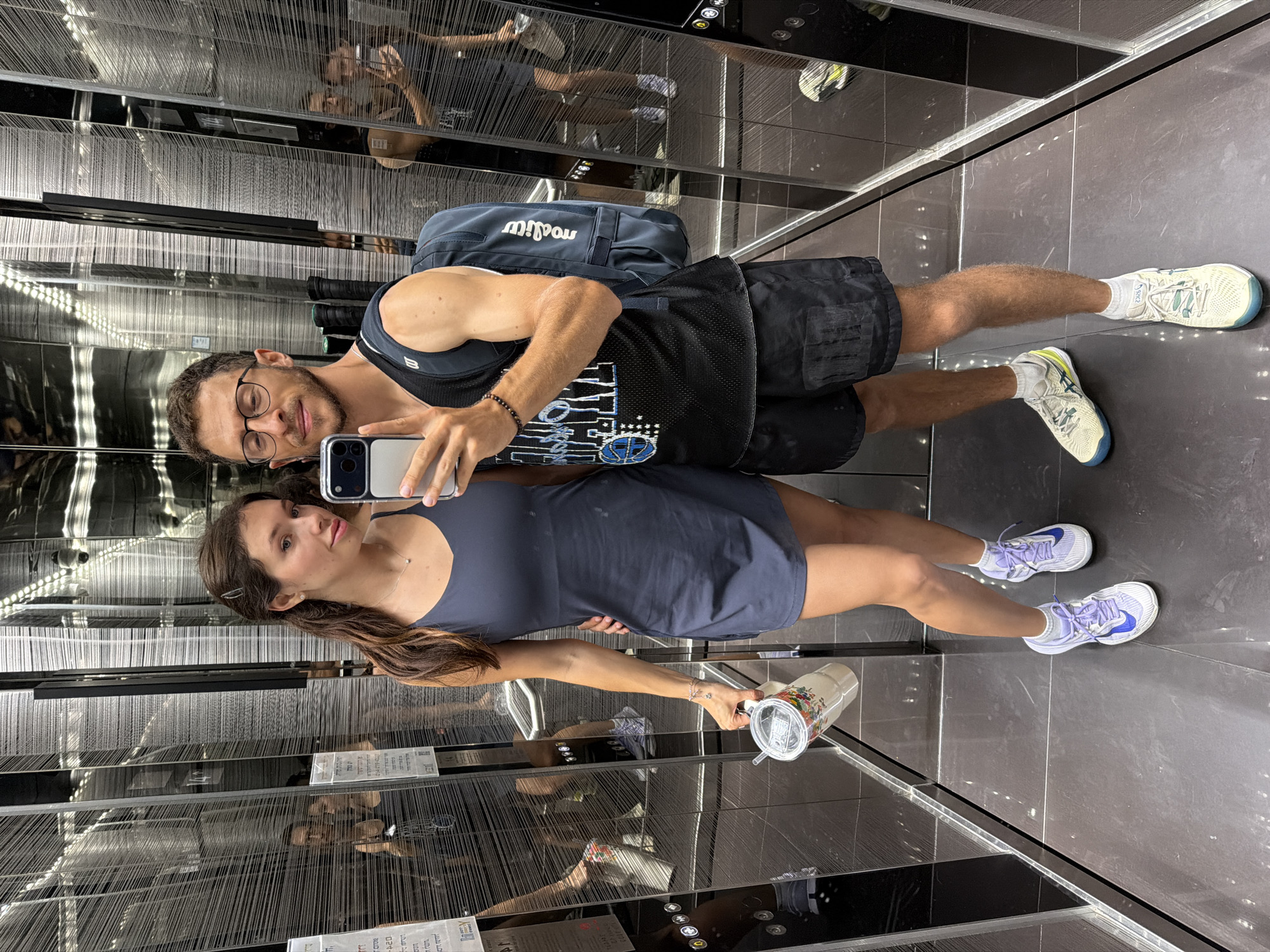 A young, athletic-looking couple is taking a selfie in a modern elevator with mirrored walls. The woman, with long brown hair, is wearing a dark blue athletic dress and white and purple sneakers. She is holding a large white cup with a straw. The man, with short brown hair and glasses, is wearing a black athletic tank top, black shorts, and white and yellow sneakers. He has a dark blue backpack on his back and is holding his phone to take the picture. The elevator walls are highly reflective, showing multiple reflections of the couple and the elevator interior, including the buttons and lighting. The floor is dark and polished.
