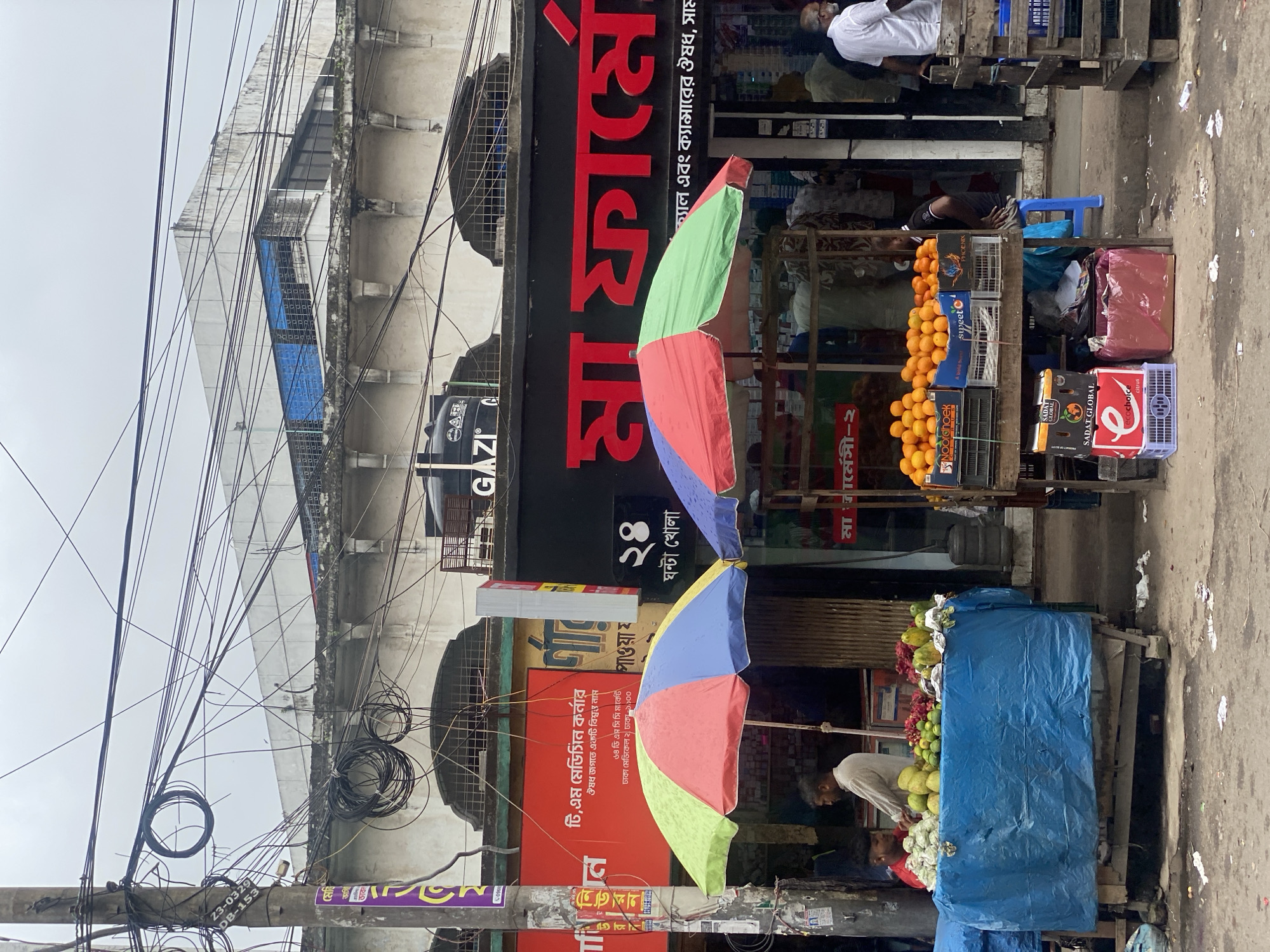 The image depicts a street scene in Dhaka, Bangladesh, characterized by a dense network of overhead electrical wires and a cloudy sky. In the foreground, a fruit stall is prominently displayed, covered with a blue tarp, with various fruits like guavas and limes arranged on top. Two vendors are visible near the stall. Adjacent to the fruit stall, a pharmacy with a red sign is operating. Further to the right, another stall selling oranges in crates is set up, with a person attending to it. The street is paved and shows signs of being a busy commercial area.