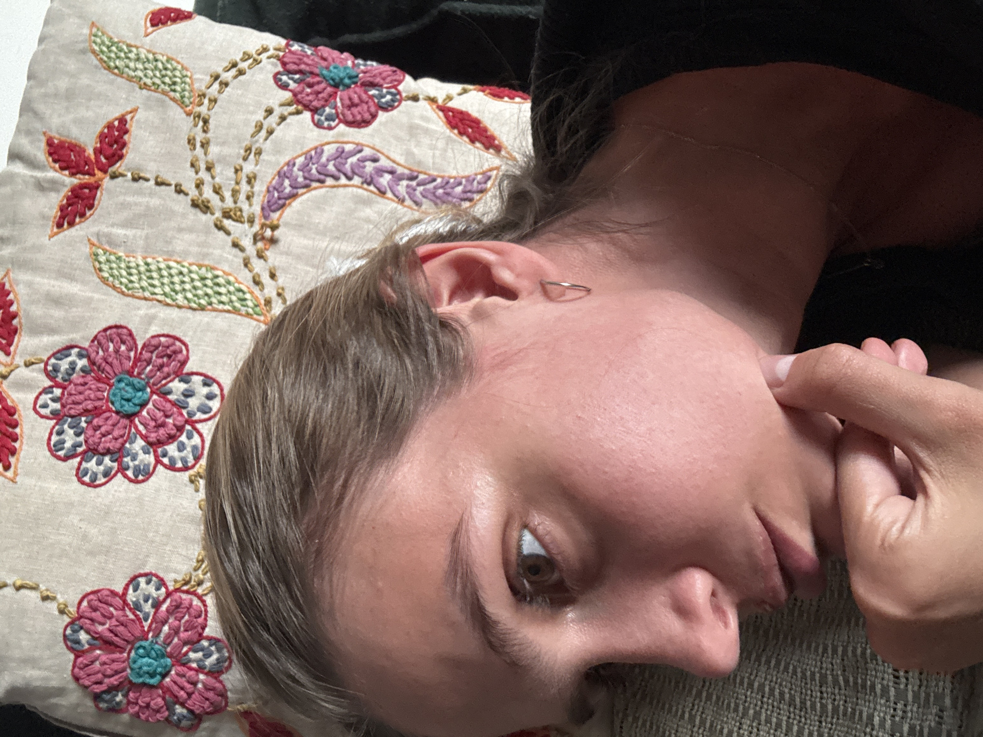 A person lies on a pillow with floral embroidery. The person's head is tilted, and they are holding their face with their hand. The location is Unknown, Unknown.