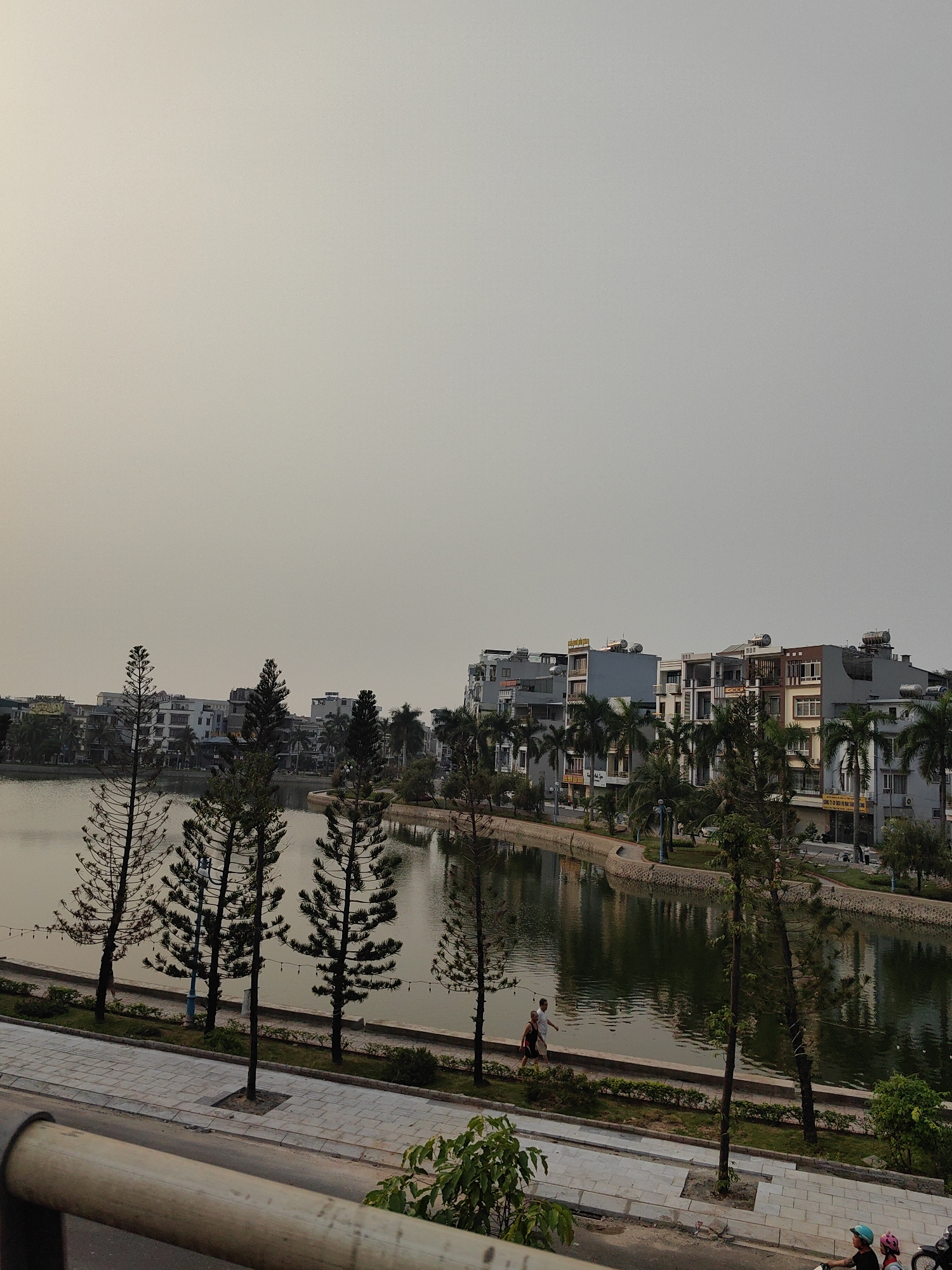 A view of the lake in Hong Gai Ward, Vietnam. The lake is surrounded by modern buildings and trees. Several pine trees line the edge of the lake. A man and a girl are walking on the paved path near the water. In the lower right corner, two children on a motorbike are wearing helmets.