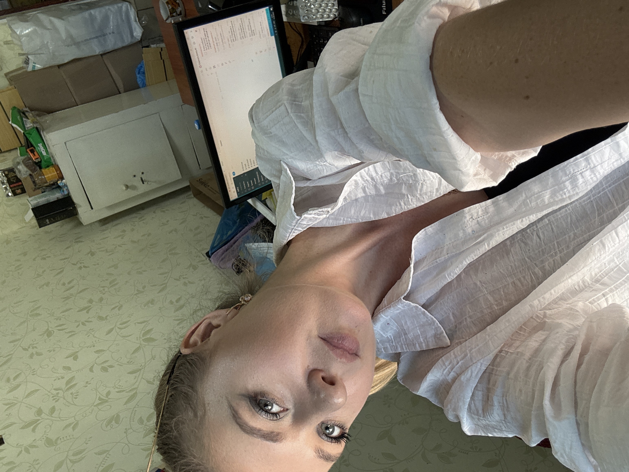A woman with blonde hair and blue eyes is taking a selfie in an office. She is wearing a white collared shirt. Behind her, there is a computer monitor, a desk, and a safe. The room is cluttered with boxes and other items. The lighting is dim, and the overall atmosphere is casual.