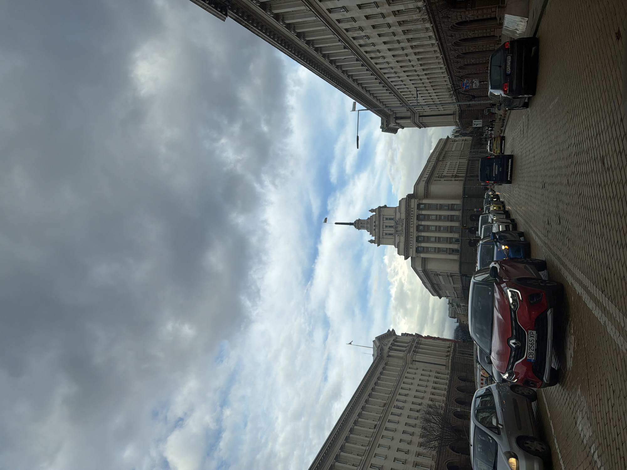 The image shows a street scene in Sofia, Bulgaria, with a line of cars on a cobblestone street facing away from the viewer. The architecture on both sides of the street is tall and imposing, with many windows and intricate details. One notable building has a tall, layered structure with a flag on top. The sky is overcast with a mix of dark and light clouds. There are no visible people in the frame. The license plates of the cars are visible.