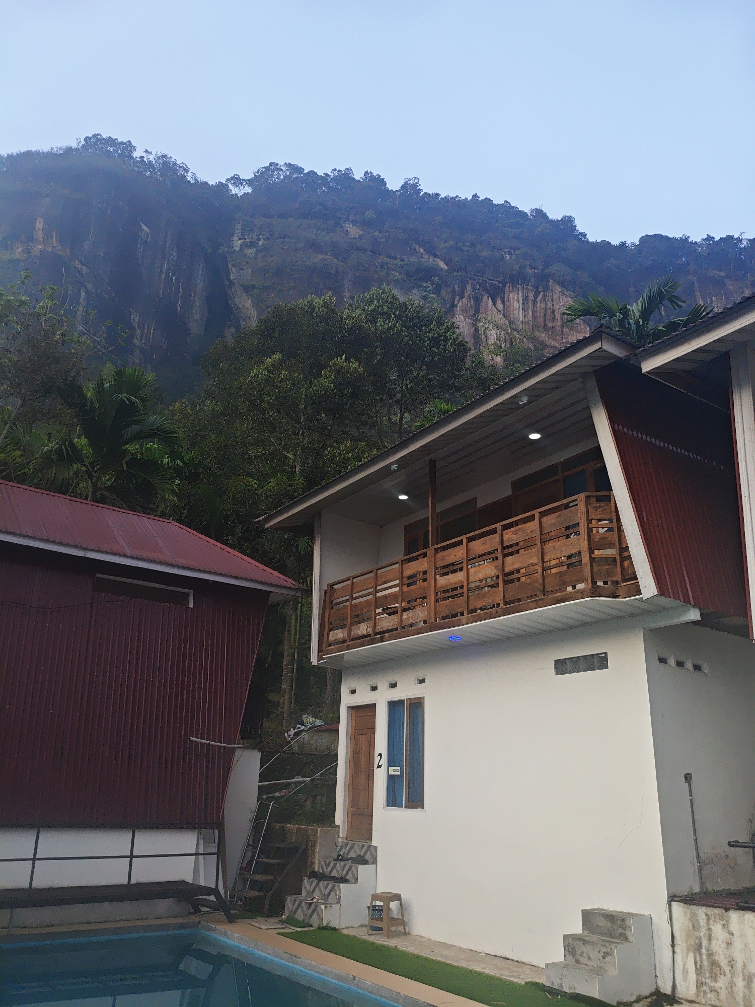 In the city of Harau, Indonesia, a two-story building with a white facade and a balcony with a wooden railing is captured against a backdrop of a steep, tree-covered cliff. A smaller, dark red corrugated metal building is situated to the left of the main structure, partially obscuring a swimming pool in the foreground. The scene is set during daylight, with overcast skies. The environment is lush and natural, with dense vegetation and rocky cliffs dominating the background. The architecture appears functional, suggesting a lodging or resort accommodation.