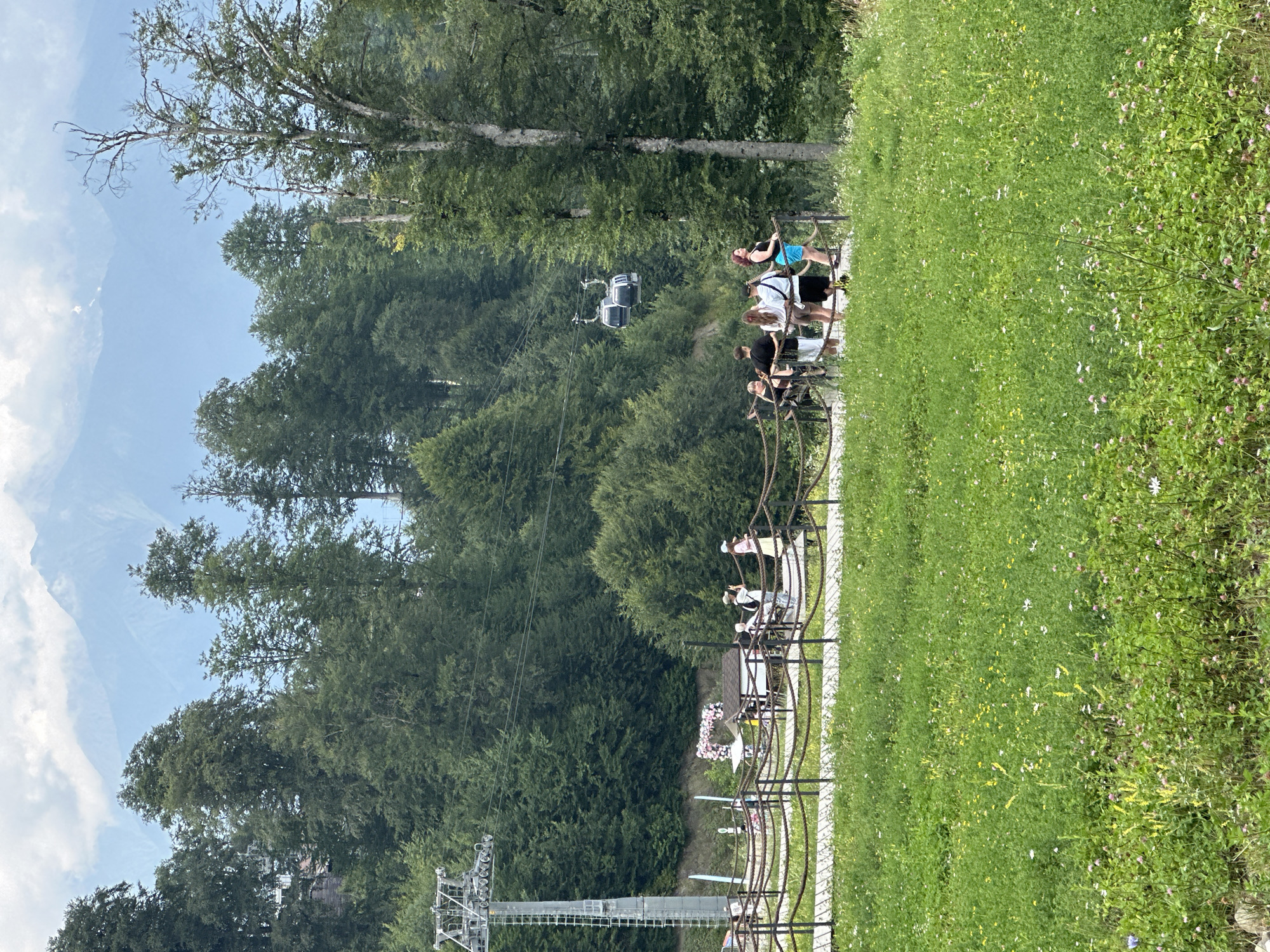 The image depicts a scenic mountain landscape with a lush green meadow in the foreground. In the mid-ground, a wooden walkway with rope railings is bordered by trees and leads to a group of people. Behind them, a cable car system is visible, with two cabins suspended in the air against a backdrop of dense green forest. Further in the distance, majestic mountains rise, their peaks partially obscured by clouds. The scene appears to be set in the popular resort area of Estosadok, Russia, known for its natural beauty and outdoor recreational activities. A small structure decorated with pink flowers is also visible near the walkway. The overall impression is one of a serene and picturesque natural environment, likely a tourist destination.