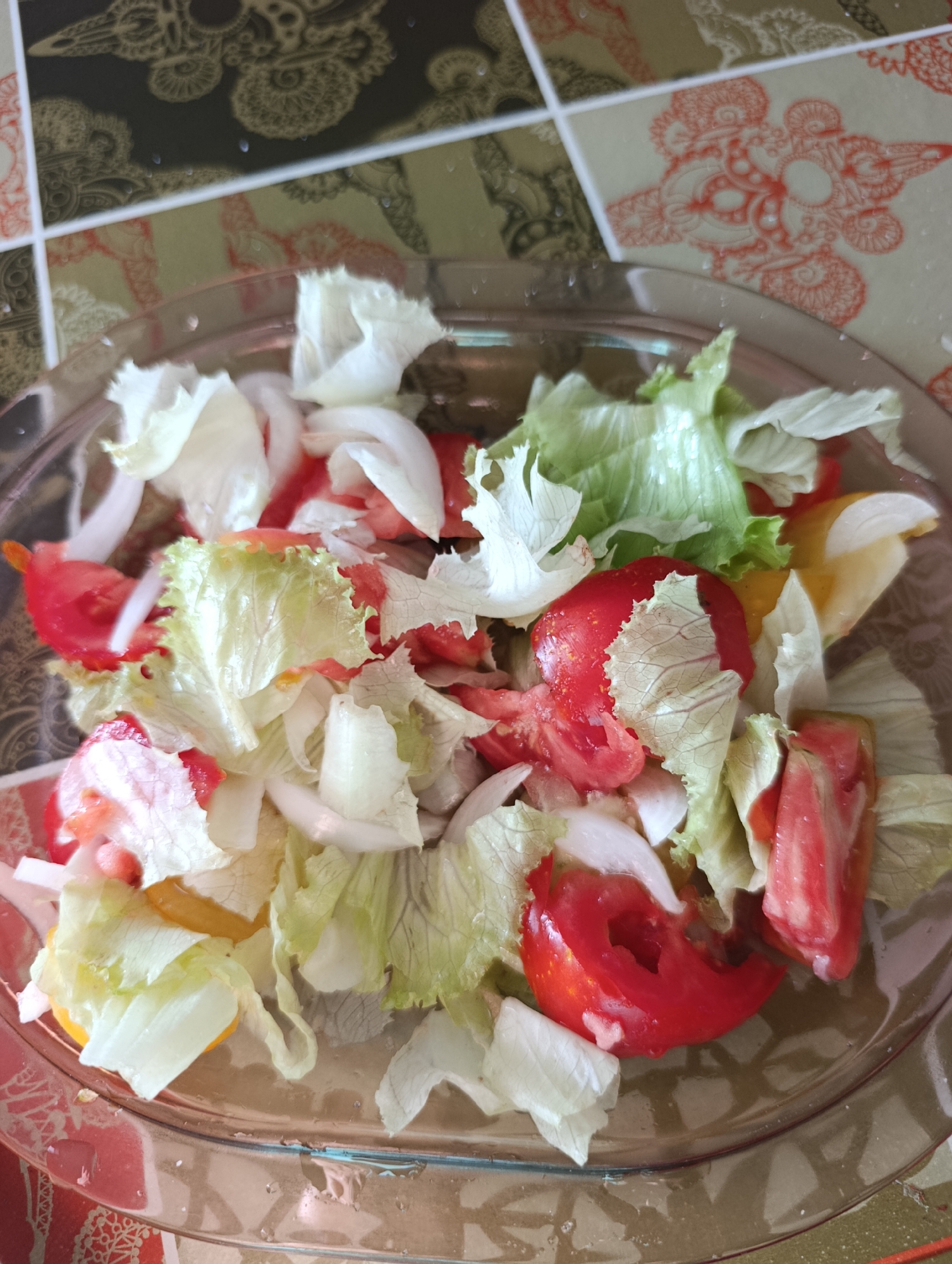 A close-up, overhead shot shows a clear glass bowl filled with a fresh salad. The salad is composed of chopped red tomatoes, white onion slices, and crisp green lettuce. The ingredients are mixed together, with some pieces of yellow bell pepper visible underneath. The salad is presented on a table with a patterned tablecloth. The tablecloth has an intricate design with shades of brown, gold, and red, possibly reflecting Portuguese tile patterns, common in Setúbal, Portugal. The lighting suggests it is daytime, likely indoors. There are no people or actions depicted.