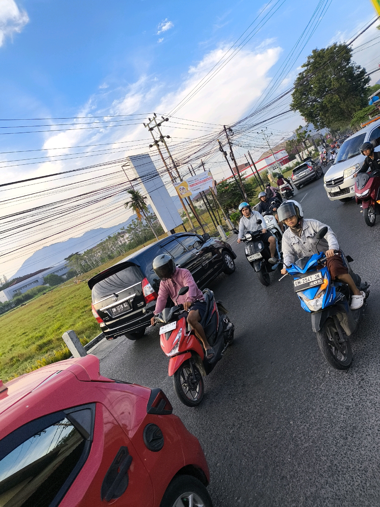 The image depicts a street scene in the city of Gorontalo, Indonesia, likely during daytime given the clear sky with scattered clouds and the quality of light. The perspective is from a low angle, looking upwards and diagonally across the road. Several vehicles are present, including a black SUV, a silver car, and multiple motorcycles. People are riding these motorcycles, most wearing helmets for safety. The road surface appears to be asphalt. On the left side of the road, there is a large grassy field bordered by a low concrete barrier. In the background, there are buildings, utility poles with numerous electrical wires crisscrossing, and a distant mountain range. The atmosphere suggests a moderate level of traffic.