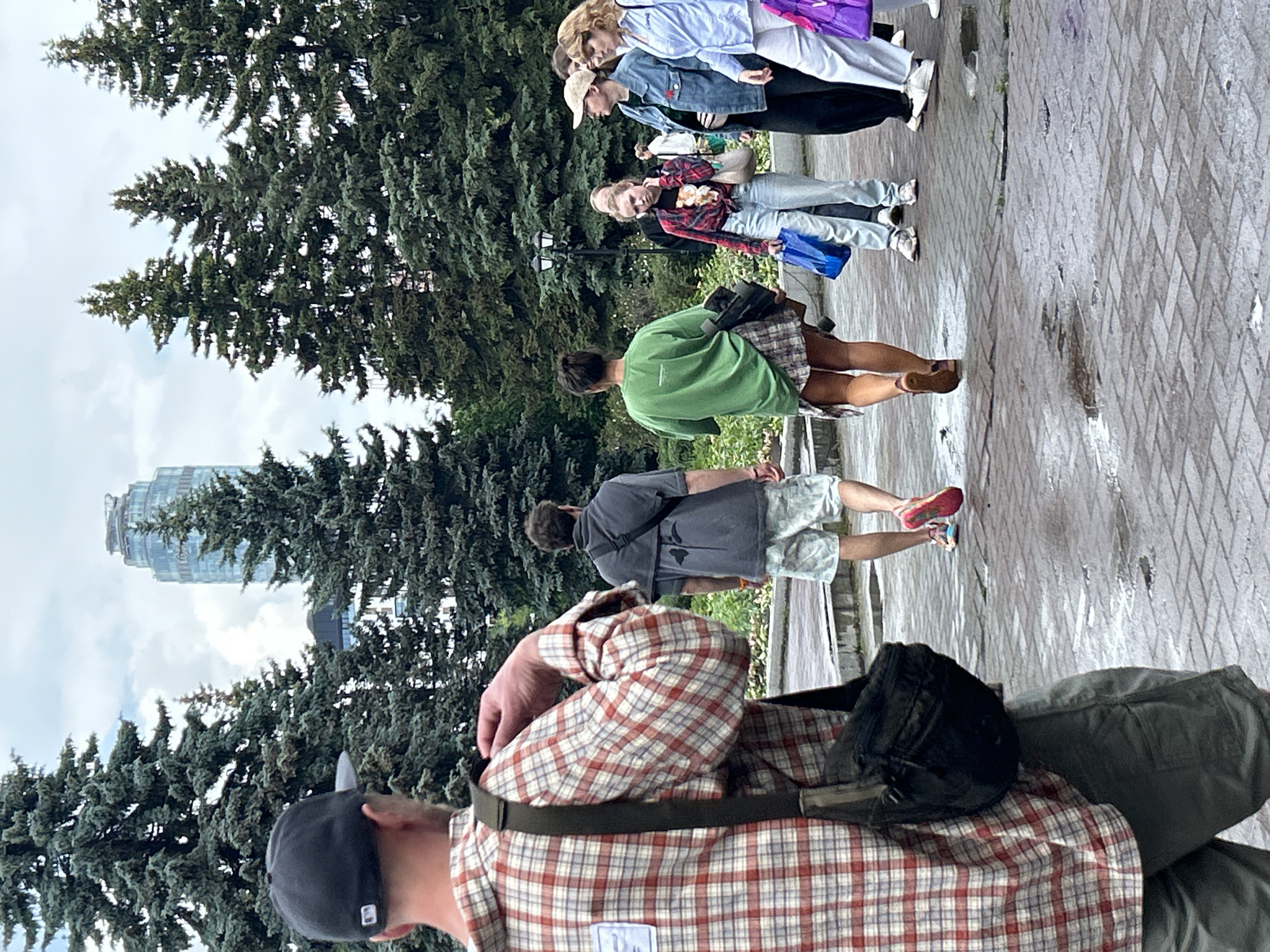 The image depicts an overcast urban park scene in Yekaterinburg, Russia, with the distinct Vysotsky Business Center skyscraper visible in the distant background. The ground is a wet, brick-paved walkway, indicating recent rain, with visible puddles. Numerous tall, dark green coniferous trees flank both sides of the pathway.
In the foreground, a man with his back to the camera wears a dark baseball cap, a red and white plaid long-sleeved shirt, and dark shorts. A black sling bag hangs across his left shoulder, and he appears to be adjusting the cuff of his right sleeve.
Further down the pathway, moving away from the viewer, are two individuals. The first is a man wearing a dark grey short-sleeved top with a black stylized design on the back, light-colored patterned shorts, and colorful running shoes (pink/red and blue). He also carries a dark sling bag. Following him is a woman, also with her back to the camera, wearing an oversized green t-shirt and a plaid skirt, with dark hair tied up.
On the right side of the image, a group of people is gathered near the pathway. A woman in a red plaid shirt tied at the waist over a black t-shirt, light blue jeans, and white sneakers holds a blue shopping bag. Next to her, a woman in a beige baseball cap, a light blue denim jacket, and dark pants wears a white fanny pack. Another woman with wavy blonde hair, a light blue long-sleeved shirt, and white pants holds a purple shopping bag. Other figures are partially visible within this group and in the background. A black street lamp with two globes stands amidst the trees in the mid-ground.