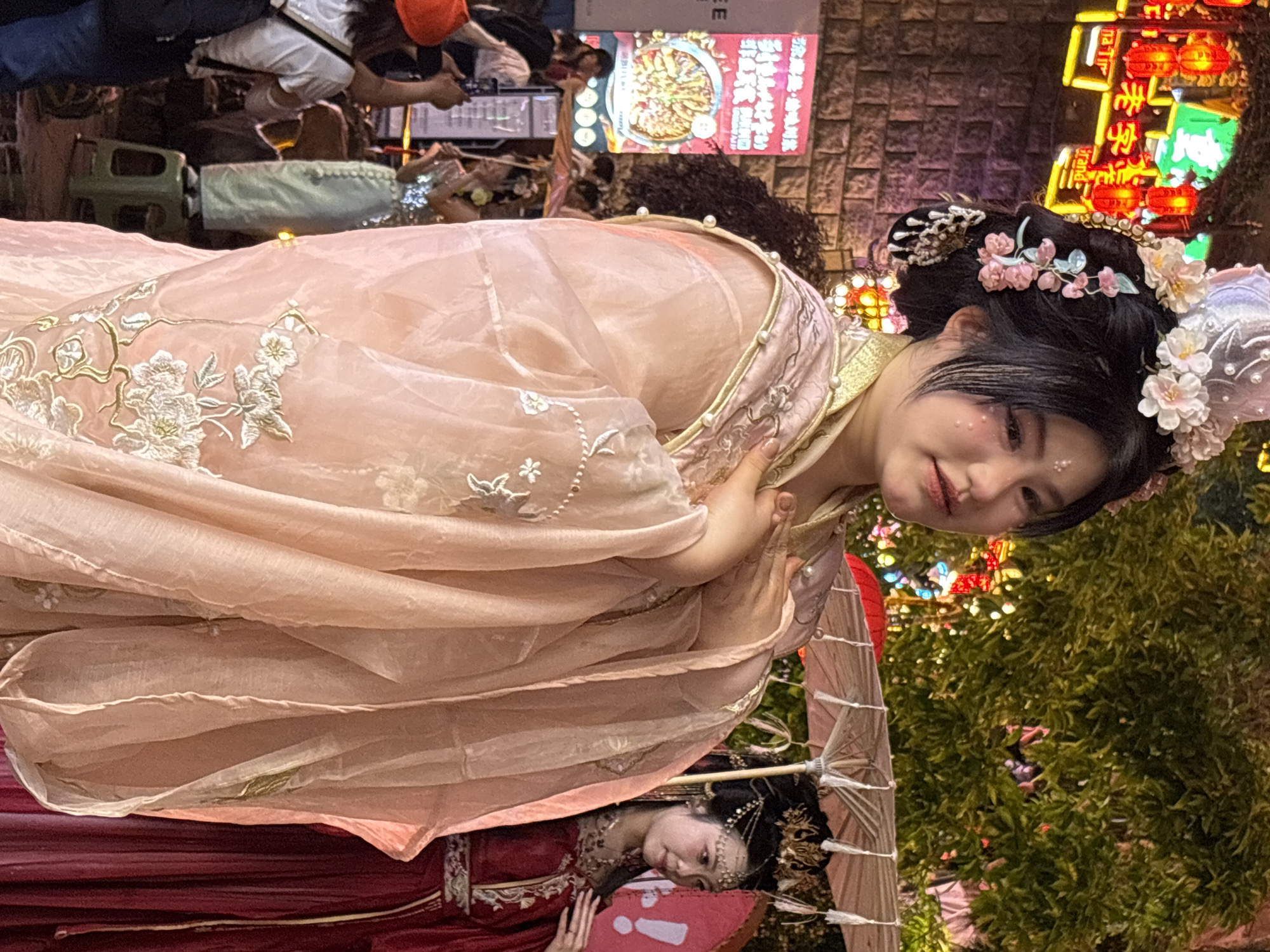 A vertical, eye-level shot features a woman in traditional Chinese clothing against an urban backdrop, likely in Yuzhong District, China. She wears a peach-colored robe with gold trim and intricate floral embroidery, her black hair adorned with floral hairpieces. Her face is turned towards the viewer, and she is smiling, with decorative sparkles on her cheek.
Below her, another woman in a red outfit is partially visible, lying down with an umbrella. People are walking in the background, and a restaurant sign is visible. The area is lit by warm, ambient lights. Green trees and shrubs are visible.