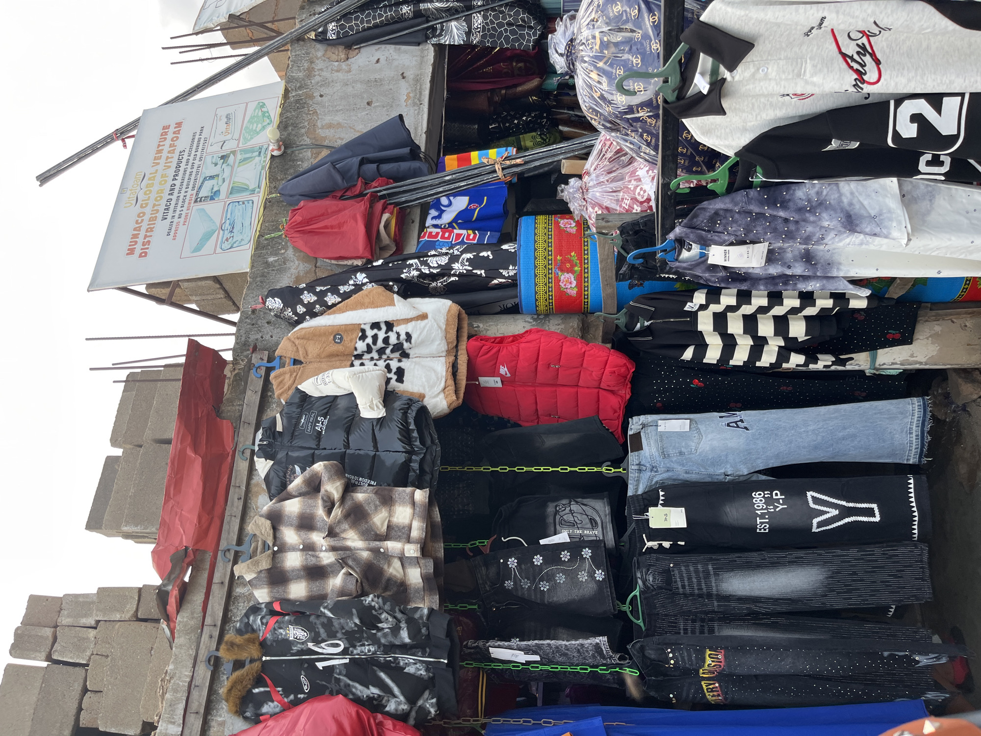 This is a medium shot, outdoor photograph taken in Bukuru, Nigeria. The image captures a street market stall displaying a variety of clothing items, predominantly outerwear like jackets and shirts, alongside some folded pants. The stall is constructed with concrete blocks and rough wooden planks, suggesting an informal setting. Hanging clothing is arranged on racks and shelves.
Notable objects include several jackets with fur-lined hoods, puffer jackets, and plaid shirts. There are also t-shirts and sweatshirts with graphic prints and text. Some of the clothing is neatly folded, while other items are displayed on hangers. On the left side of the frame, a large white sign with red and blue lettering advertises "MUNACO GLOBAL VENTURE DISTRIBUTOR OF VITAFOAM" and lists an address and phone numbers, providing a specific location clue.
The scene appears to be during daylight, with the sky visible at the top of the image and a bright, overcast lighting. There are no people directly visible in the frame, but the display of goods indicates a commercial activity, likely a retail sale. The overall impression is of a busy, functional market environment. Visible text includes brand names and descriptive phrases on the clothing and the prominent advertisement sign.