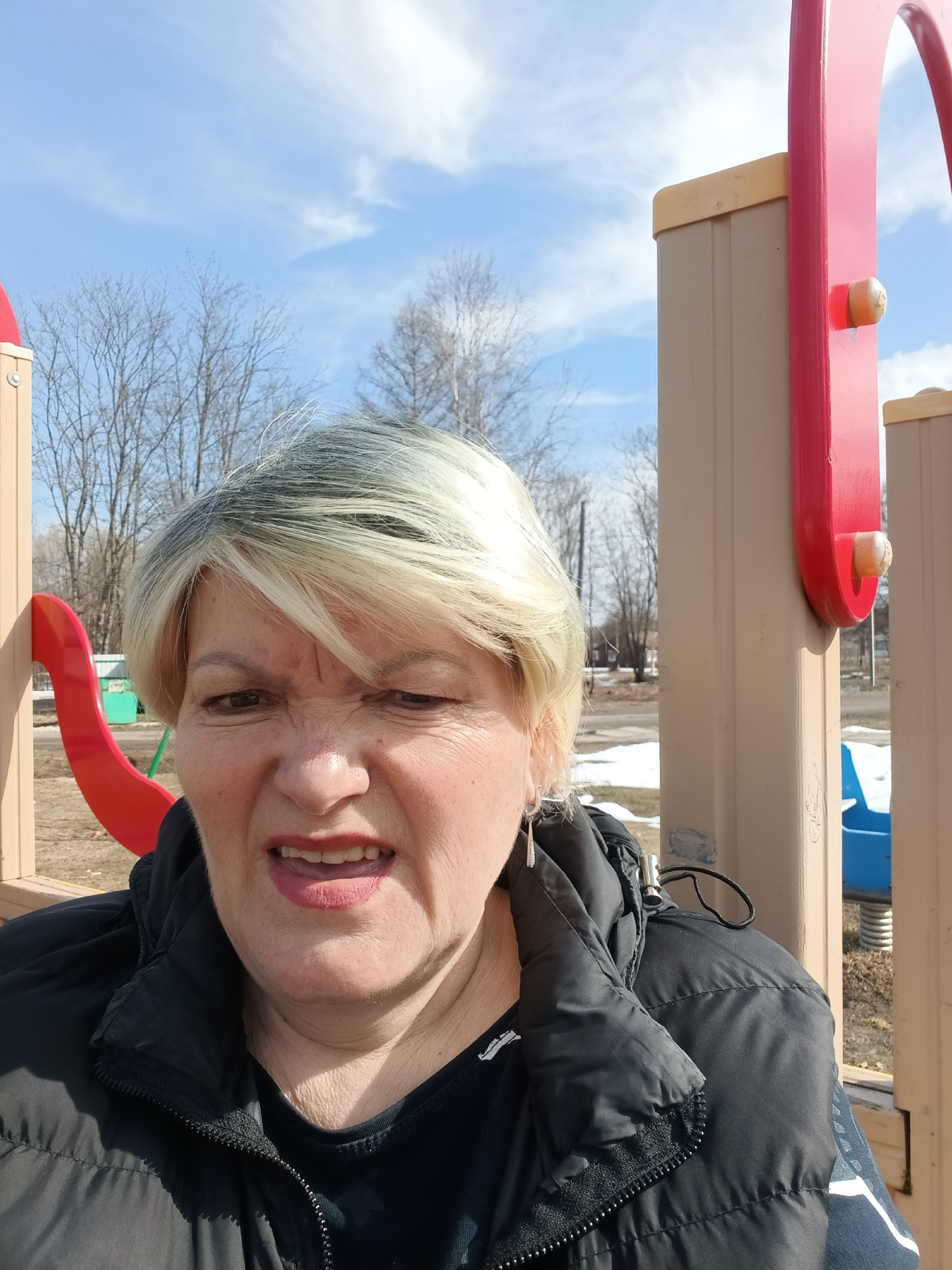 The image shows a woman in a black puffer jacket at a playground. The playground equipment is painted in red, blue, and tan colors. Bare trees are visible in the background, along with patches of snow. The sky is blue with scattered clouds. The location is Красный Маяк, Russia.