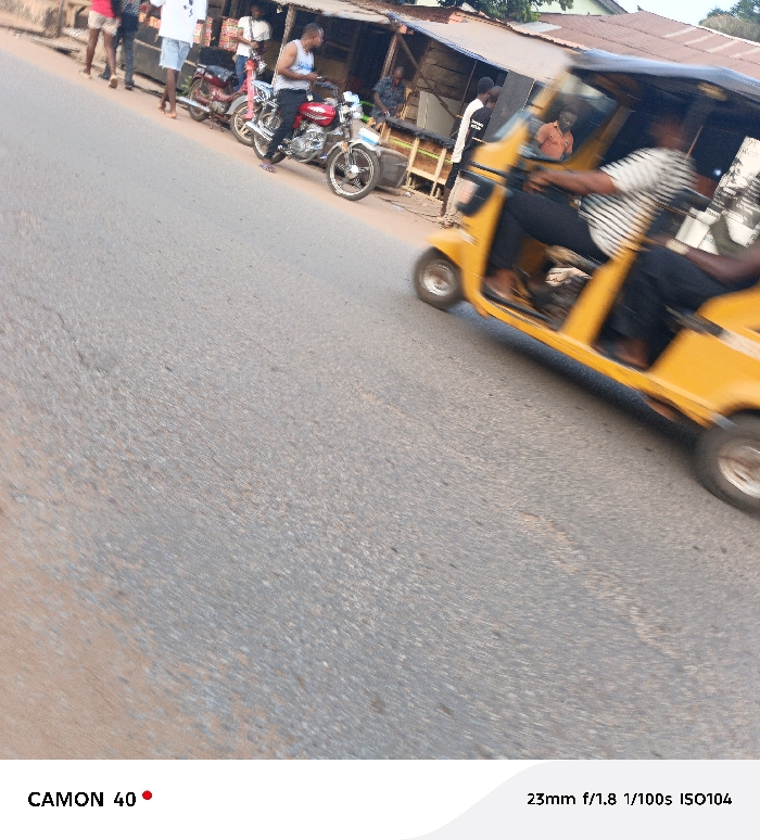 A blurred yellow auto rickshaw is captured in motion, moving from left to right. The driver, wearing a striped shirt, appears partially visible. In the background, people are standing by parked motorcycles near a building. The road occupies a significant portion of the foreground and extends towards the horizon. The photograph is taken in daylight with details of the street visible.