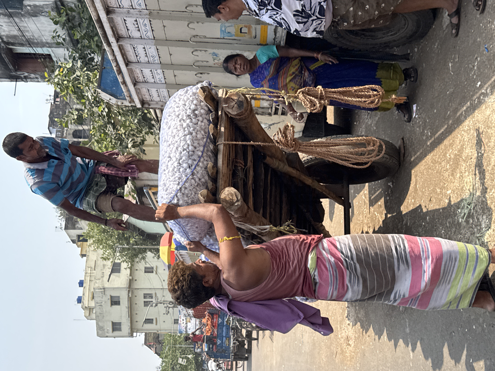 In Kolkata, India, a group of people are unloading a large sack filled with white cotton-like material from a wooden cart. One man, wearing a blue and striped shirt, is on the left side of the cart. Another man, wearing a pink tank top and a sarong, is lifting the sack, and he has a purple cloth draped on his shoulder. Two other people are next to a truck behind the cart, one man wearing a patterned shirt and one woman wearing a blue sari. The environment includes buildings, trees, and other vehicles on the street, suggesting a busy urban setting. The cart is constructed of wood and rope and has small tires.