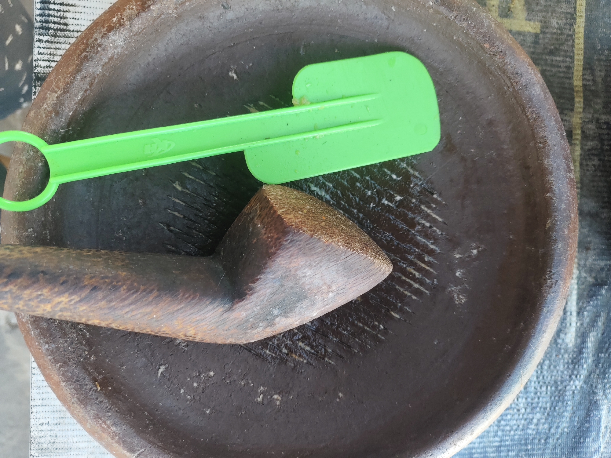 A mortar and pestle are positioned on a patterned cloth, likely in an outdoor setting in Bangsalsari, Indonesia. The mortar, made of dark brown clay, is concave with visible striations indicating use. A wooden pestle with a smooth, rounded handle and a beveled striking end rests within the mortar. A bright green plastic spatula with a notched edge is placed on top of the mortar, with its handle extending leftwards and its blade angled towards the right. The spatula has visible residue, suggesting recent use. The cloth beneath the mortar has a rough texture and features a repeating pattern of thin black and white lines, along with thicker yellow and white stripes. The overall scene suggests food preparation or processing.