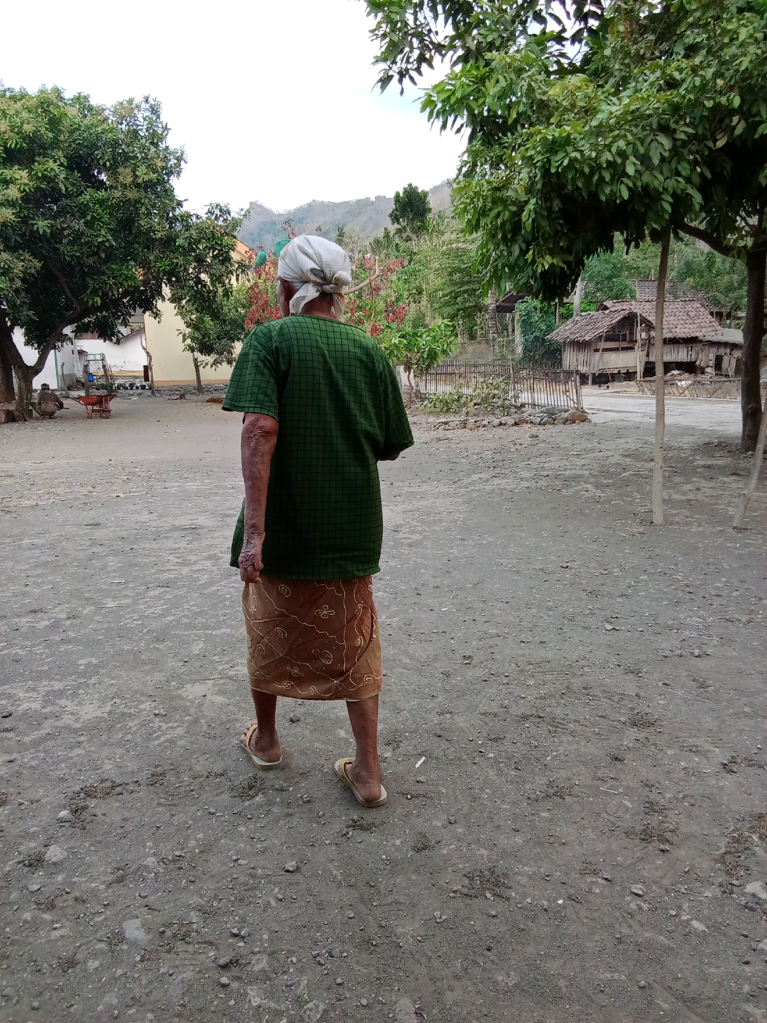 A woman wearing a green checkered short-sleeved shirt, a brown patterned sarong, and light-colored flip-flops walks away from the camera on a dirt path in a rural area. She has a white headscarf tied around her head. Her arms appear weathered. In the background, there are trees, a simple wooden structure with a thatched roof, a fence, and some buildings. A wheelbarrow is visible to the left of the image. The environment suggests a village setting in Trebungan, Indonesia.