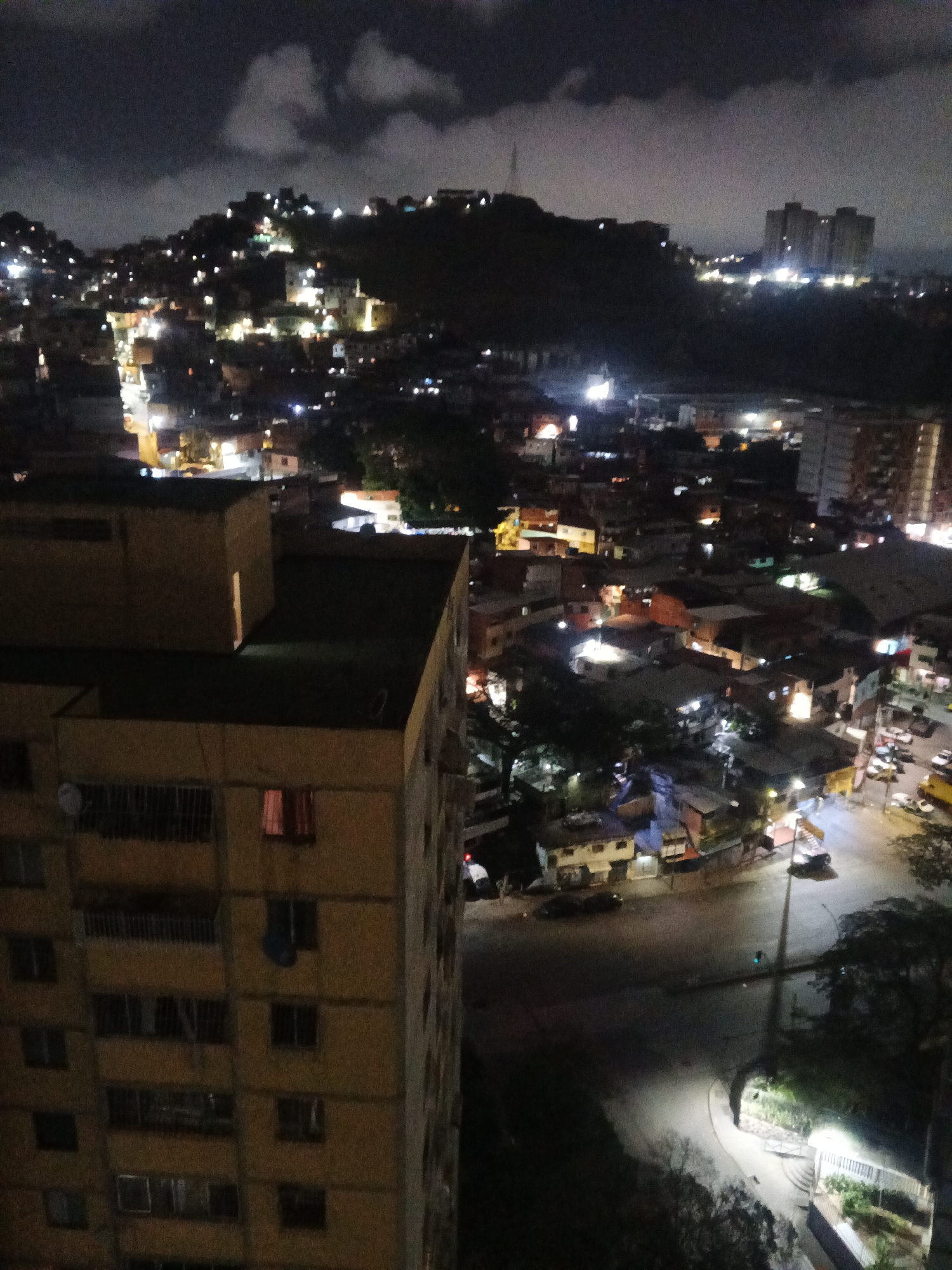 The image captures a night view over Caracas, Venezuela, likely from a high vantage point within the city. In the foreground, a tall, yellow-ish apartment building dominates the left side of the frame, with several visible windows and at least one item of clothing hanging from a balcony. The right side of the image shows a section of the city, with streets illuminated by streetlights and the lights of homes. There are several cars parked on the side of the road. In the background, a hill covered in buildings extends across the horizon, also illuminated by the lights of homes and the city. Higher up, some taller buildings stand out, bathed in light. The night sky is dark, with scattered clouds.