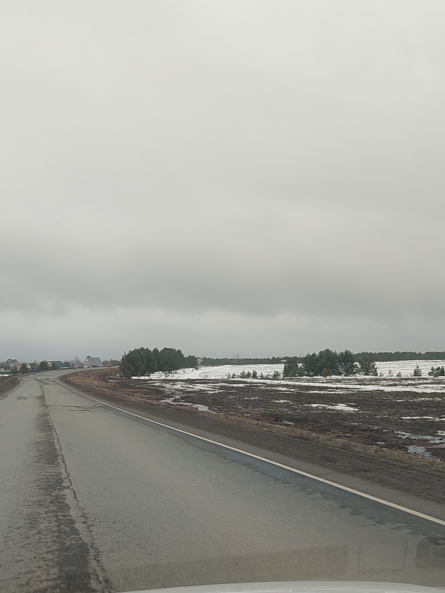A photograph depicts a grey, overcast day in what appears to be a rural area. A paved road curves gently to the left, taking up the lower left of the frame. The road surface is wet and shows signs of wear, with visible cracks and patches. To the right of the road is a snow-dusted field, interspersed with patches of brown vegetation and scattered small trees. In the distance, a faint outline of structures can be observed near a treeline. The sky is uniformly grey and occupies the majority of the upper portion of the photograph, with no visible sun or breaks in the cloud cover. The setting is possibly near Povarenki, Russia, based on context.