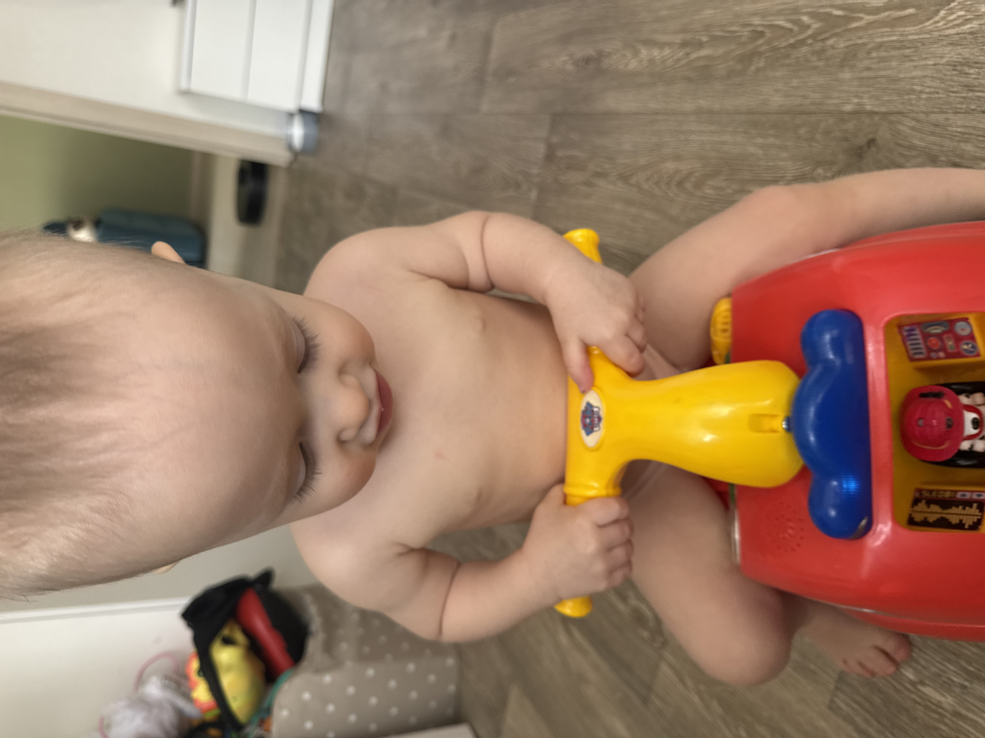A baby is seated on the floor, playing with a red and yellow toy. The baby appears to be indoors on a wooden floor, with a door visible in the background. Additional toys are visible in a corner. No information available to connect the photo to Chaikovsky, Russia.