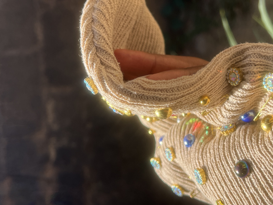 The image presents a close-up, high-detail view of a light-beige, ribbed knit garment, possibly a sleeve or a portion of a larger textile item. The fabric is heavily embellished with an assortment of decorative elements, including numerous gold-toned dome-shaped studs, various iridescent and faceted gemstones in gold settings (some displaying an aqua hue, others a rainbow spectrum), dark blue teardrop-shaped beads, and several gold-toned safety pins. A person's hand or wrist, characterized by dark-toned skin, is visible within or adjacent to the garment, suggesting the item is being worn or held. The garment is slightly creased and folded, indicating movement or a natural drape. The scene is illuminated by strong, direct sunlight, creating bright reflections on the metallic studs and iridescent gemstones. The background is out of focus and appears dark brown to black, with a blurred green element discernible in the lower right quadrant, suggesting an outdoor environment. This scene is located in Shen, Nigeria.