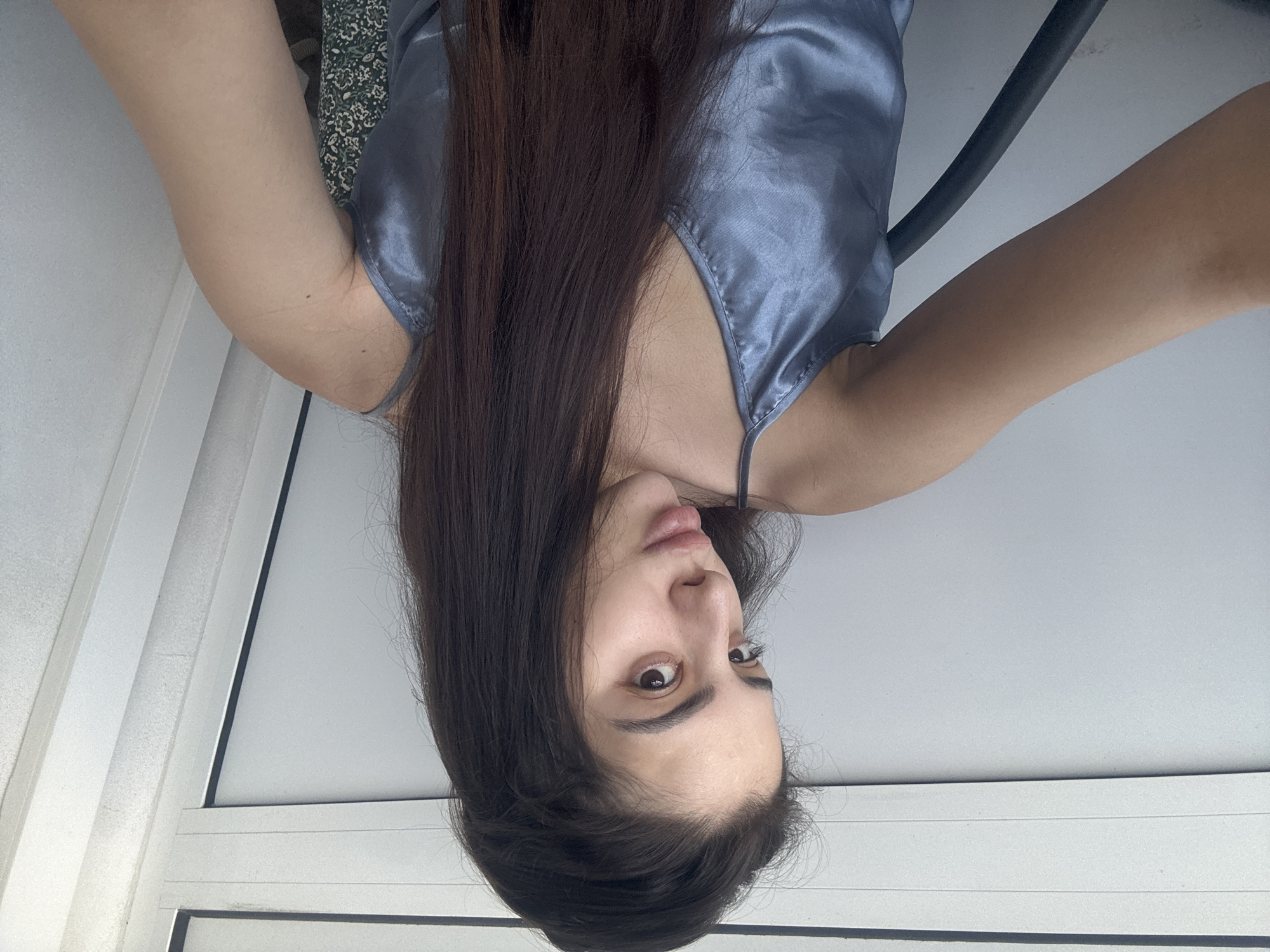 The image features a young woman capturing a selfie from an overhead perspective, appearing upside down in the frame. She has long, dark brown hair that falls downwards, partially covering her shoulders and chest. She is wearing a light blue or grey silky camisole with thin spaghetti straps. Her face is turned towards the camera, displaying dark eyes, a slight pout on her lips, and a curious or playful expression. Her arms are raised, framing her upper body.
The background is a bright, plain white or light grey, suggesting an indoor setting with ample ambient light. White linear elements, possibly window frames or blinds, are visible on the left side, further indicating an interior space. A curved black object, which could be part of furniture or a railing, is present near her right arm. In the upper left corner (when viewed in original orientation), a small section of green and white patterned fabric is visible, possibly part of bedding or a cushion.
The primary activity is posing for a self-portrait. There are no overt emotional expressions beyond a direct gaze and slight pout. No specific time of day or weather can be determined from the image. No text is visible in the media file. The provided location context is Plovdiv, Bulgaria.