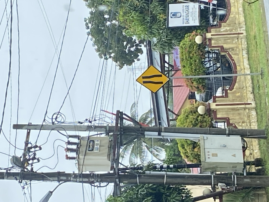 The image shows utility poles covered in wires, with electrical equipment attached. A yellow road sign with two horizontal black bars is visible. Beyond the poles are trees and a building. The building has a sign that reads "Bhayangkari." Green bushes and a red roof are visible, along with a partially obscured car. The sky is overcast.