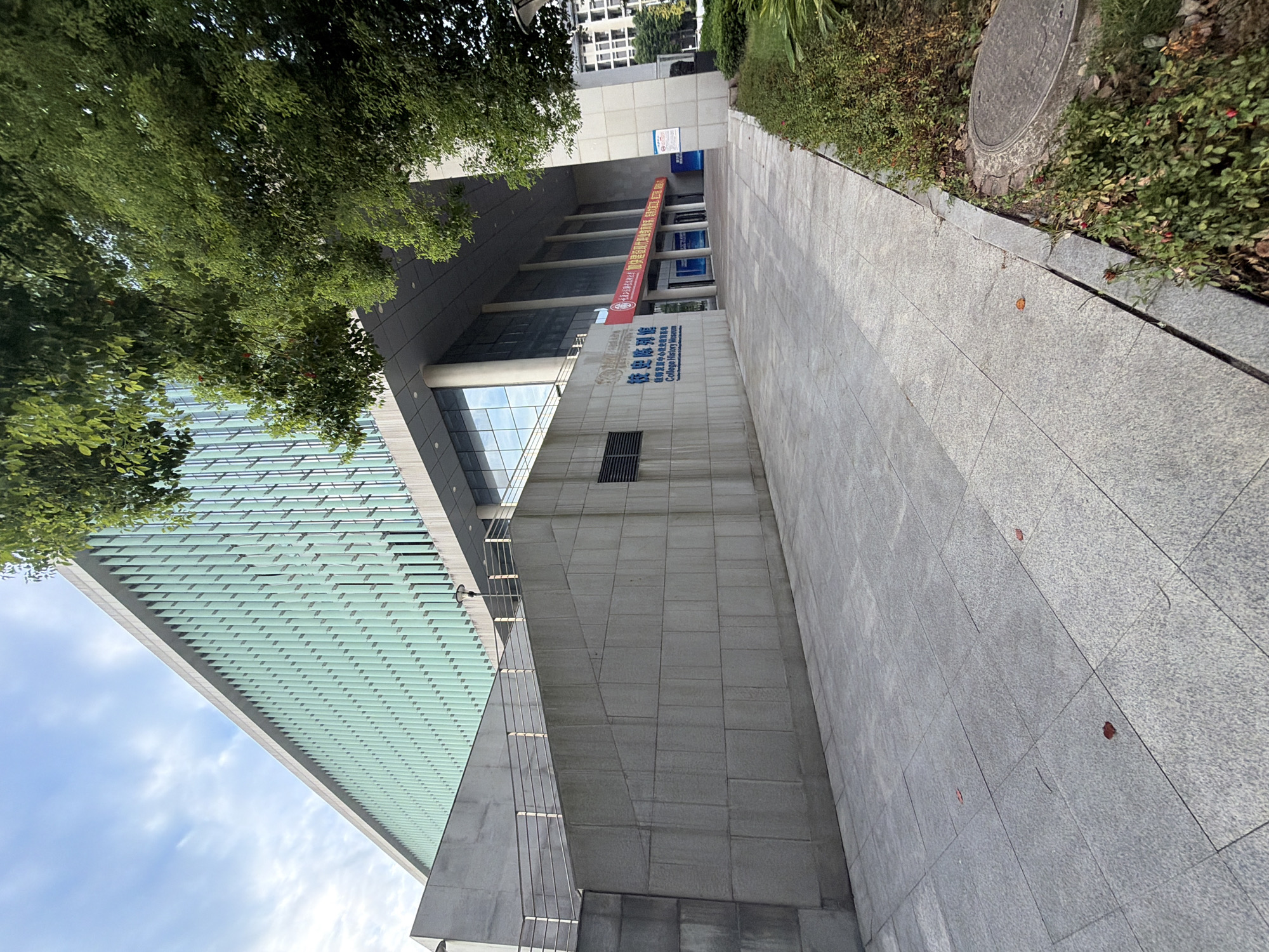 The image shows the exterior of the College History Museum in Jiangjin, China. The building features a modern architectural design with a light green, slatted facade on the upper levels and a grey stone base. A paved walkway leads to the entrance, flanked by green trees and shrubs. A red banner with Chinese characters is visible above the entrance, and signage in both Chinese and English identifies the building as the "College History Museum". The sky is partly cloudy. The image is taken from a low angle, looking upwards towards the building and the sky.