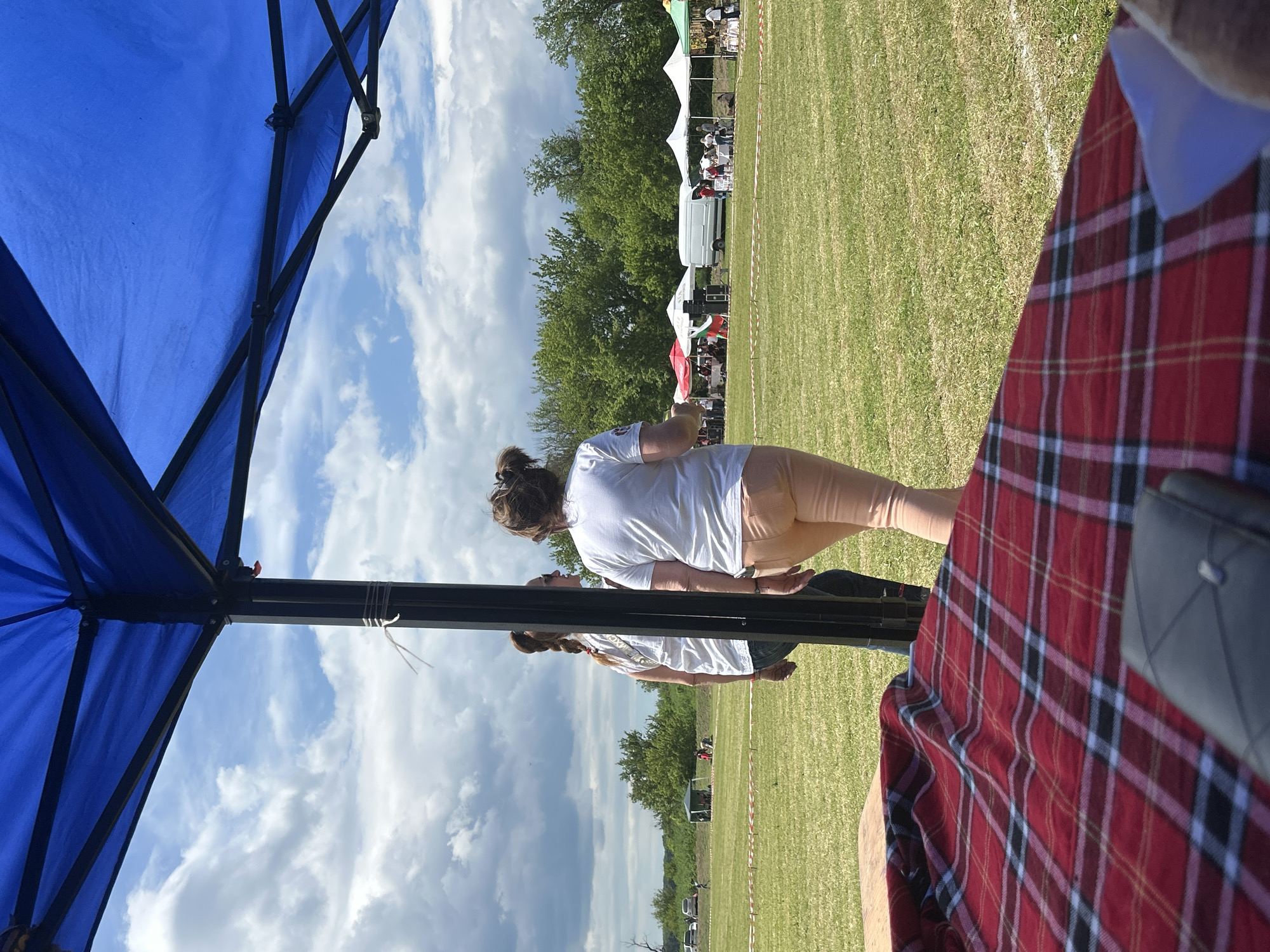 The image, seemingly taken from a low angle under a blue tent, depicts a scene in what appears to be a grassy field in Branishte, Bulgaria. In the foreground, the edge of a red and black plaid blanket is visible. A woman in a white shirt and tan pants is walking towards the center of the frame, partially obscured by a tent pole. In the background, a series of white tents are set up across the field, and trees line the horizon. A few people can be seen gathered around the tents. The sky is visible in the upper left of the frame with some puffy white clouds.