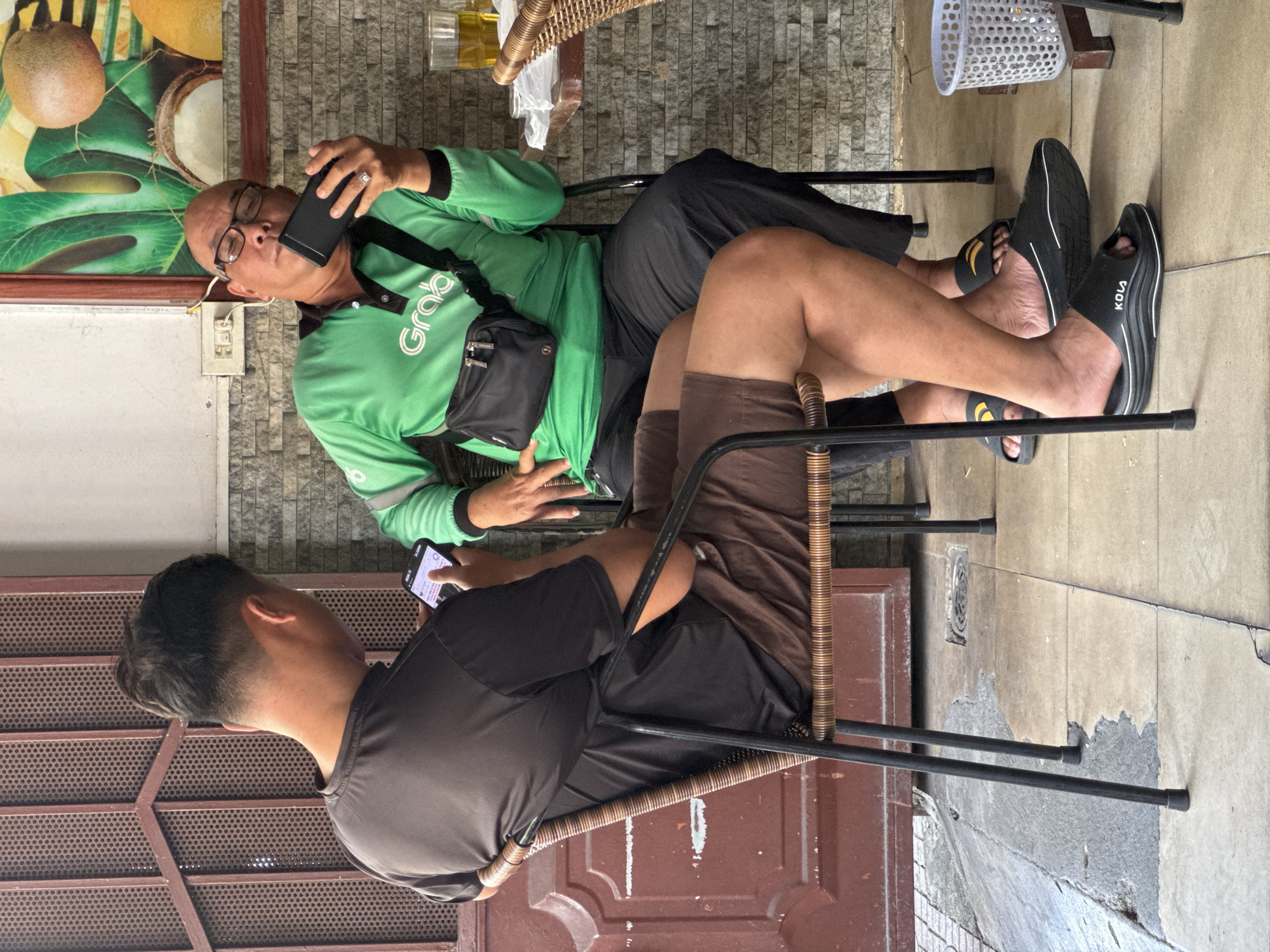 Two men are seated at a table outside a building in Tay Nha Trang Ward, Vietnam. The man on the left is wearing a dark t-shirt and brown shorts, and is looking at his phone. The man on the right is wearing a green long-sleeved shirt with the word "Grab" printed on it and black pants. He is wearing glasses and holding a black phone to his ear, appearing to be in a conversation or listening to audio. He also has a black fanny pack worn across his chest. Both men are wearing sandals. The environment includes a tiled floor, a brick wall in the background, a doorway with a perforated metal screen, and a table with a glass of amber liquid and a white basket on it. The overall scene suggests a casual outdoor setting, possibly a cafe or eatery.