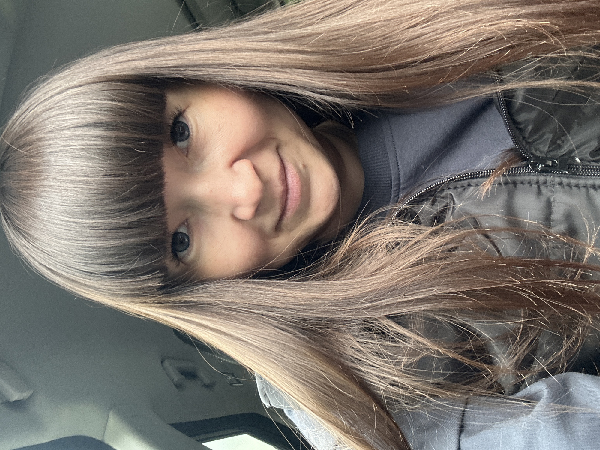 A woman with brown hair and bangs is smiling at the camera. She is wearing a gray shirt and a black jacket. The photo was taken indoors. There is no discernible evidence to indicate this picture was taken in Старые Кены, Russia.