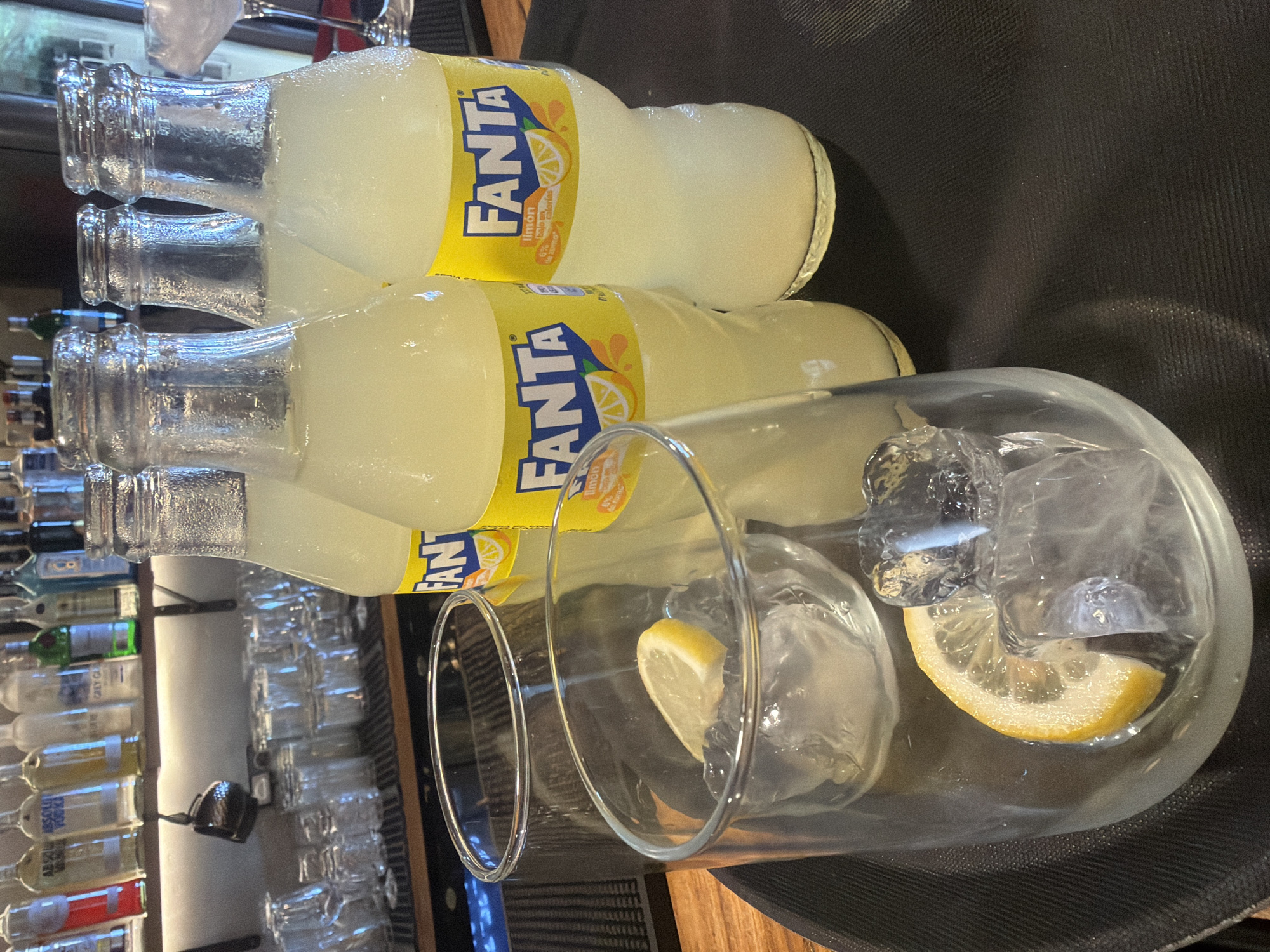 A low-angle, close-up shot captures a tray on a wooden surface, likely a bar, with four bottles of Fanta Limón and two glasses. The bottles, partially filled with a pale yellow liquid, are lined up towards the right of the frame. The front two bottles display their yellow labels with the Fanta logo and imagery of lemons. One glass in the foreground contains ice cubes and a slice of lemon, with another empty glass positioned slightly behind it. In the background, shelves are stocked with various bottles, and some glassware is visible. The overall lighting suggests an indoor setting, possibly during the day. The image does not depict any people.
