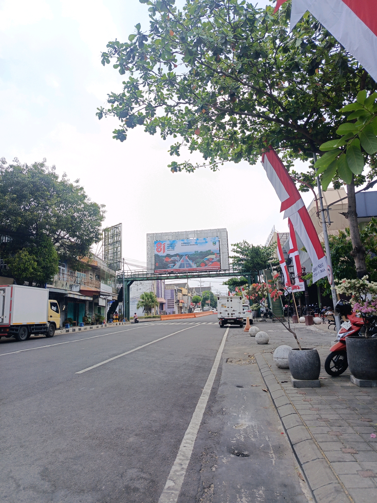 A street scene in Magetan, Indonesia, shows a road with a white lane marking. A yellow truck is parked on the left side of the road, and across the street are buildings with balconies and a large billboard in the distance. Indonesian flags are hanging from poles on the right side of the street, near potted plants and a motorcycle. The sky is overcast, and trees with green leaves are visible throughout the scene. The pavement on the right is made of brick.