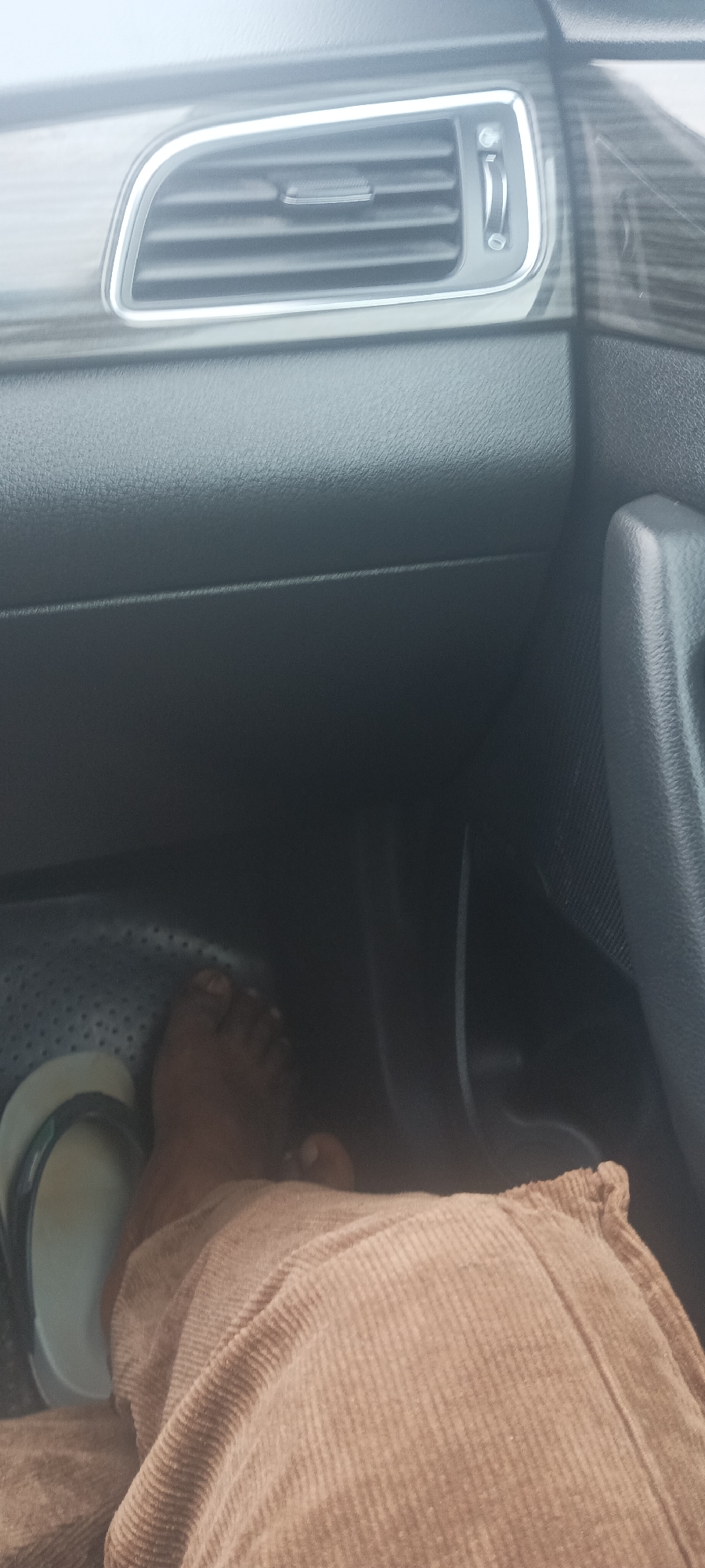 The image shows the interior of a car, specifically the passenger side footwell. A dark-skinned foot is visible, partially obscured by tan corduroy trousers. A sandal is on the foot. The car's dashboard, air vent, and door handle are also visible. The location is not discernible.