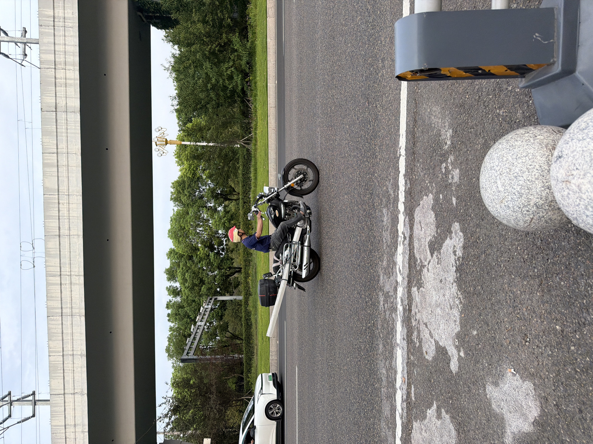 A man wearing a red helmet and a blue shirt is riding a black motorcycle on a paved road. He is carrying a long, white pole horizontally behind him, secured to the motorcycle. A white car is visible to the left of the motorcycle, also on the road. In the background, there is a large concrete overpass, trees, and utility poles. The scene appears to be in an urban or suburban area with overhead power lines and a road infrastructure. The road surface has white lane markings and some patches of lighter colored asphalt. There are also two large, spherical decorative stones and a grey traffic barrier in the foreground.