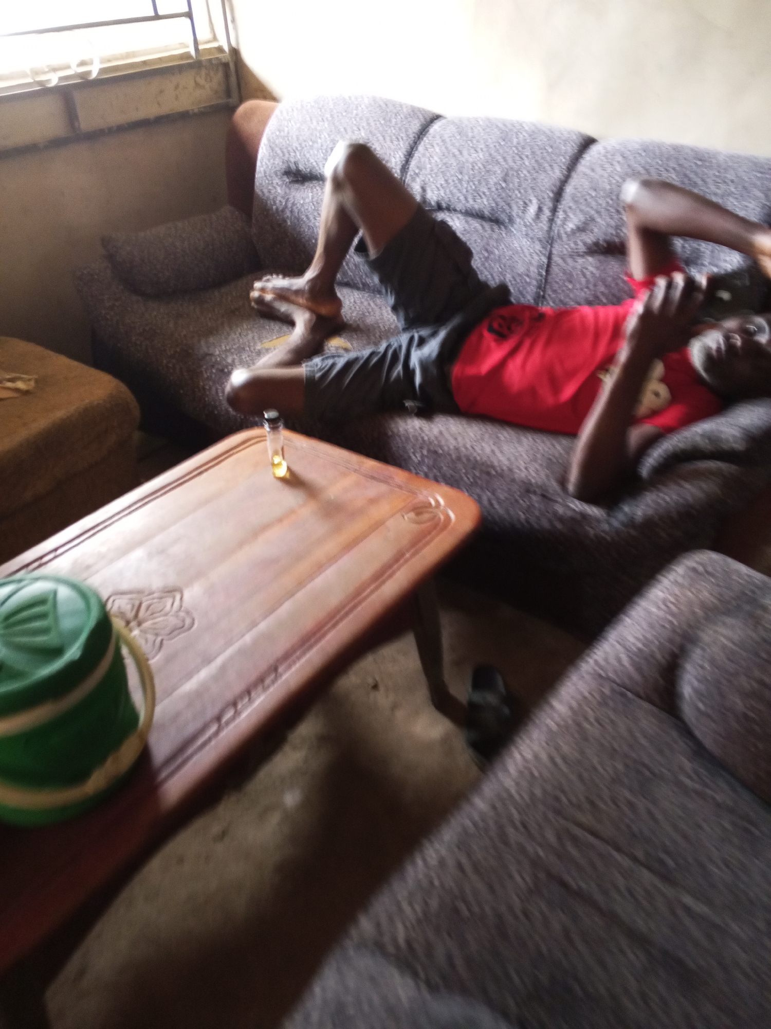 A man is reclined on a grey, patterned couch in what appears to be an indoor setting in Adonkolo, Nigeria. He is wearing a red t-shirt and dark shorts. He holds a mobile phone to his right ear and looks up. A wooden table is in the foreground, on which sits a small, clear vial containing yellow liquid and a green-and-white striped plastic bucket. The surrounding environment suggests a simple living space, with a window visible in the upper left corner. A single black shoe is visible on the floor under the table.