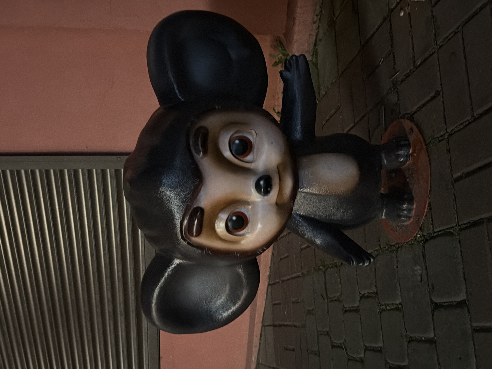 A statue of a cartoon character, Cheburashka, stands on a cobblestone pathway in front of a pink building. The statue is dark brown with large ears, a tan face, and black eyes and nose. It is wearing a light tan chest piece. The character's right arm is extended to the right. The building behind the statue has a dark, corrugated metal door or shutter. The scene appears to be outdoors, possibly in a public space. The location is identified as Kislovodsk, Russia.