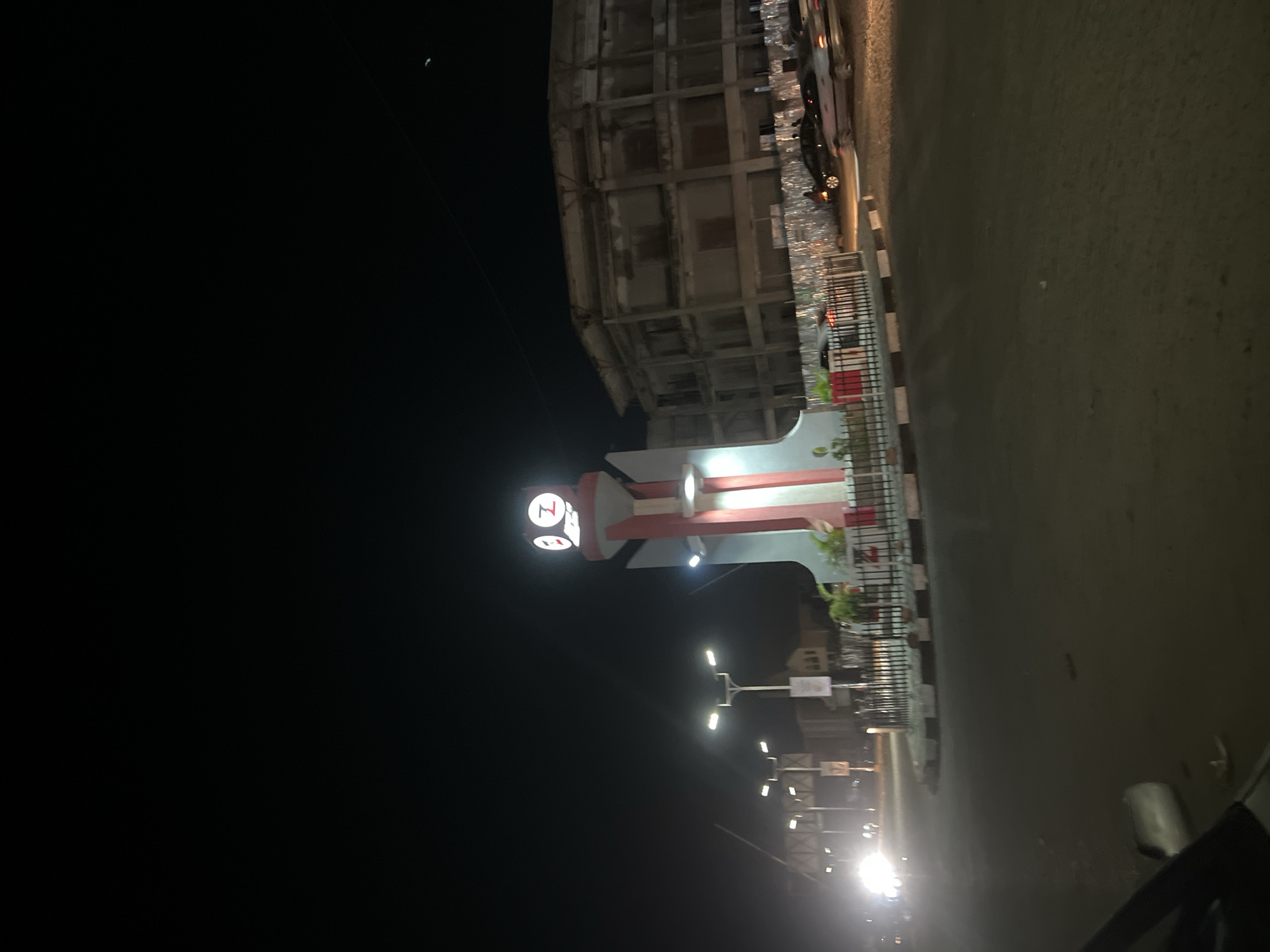 This image depicts a nighttime scene at the Zaria Road Roundabout in Jos, Plateau State, Nigeria. A prominent, illuminated clock tower stands centrally within the roundabout. The tower features a multi-tiered structure with a wide, white base tapering upwards, accented by vertical reddish-orange panels. The clock face at the top is brightly lit and displays a distinctive stylized "Z" symbol where numbers would typically be. The base of the tower is surrounded by a low, dark fence enclosing a small garden area.
To the right of the clock tower, a multi-story concrete building appears unfinished or abandoned, characterized by exposed structural elements and an absence of clear windows. This building is partially enclosed by a corrugated metal construction fence at ground level. The tarmac road of the roundabout is visible, with several vehicles present; one vehicle with bright headlights is approaching from the left, while other cars are seen on the right side of the road near the unfinished building. Multiple streetlights with bright white illumination are positioned around the roundabout, casting light on the scene. The night sky is dark, with a faint crescent moon visible in the upper right corner of the frame. No distinct human figures are clearly discernible.