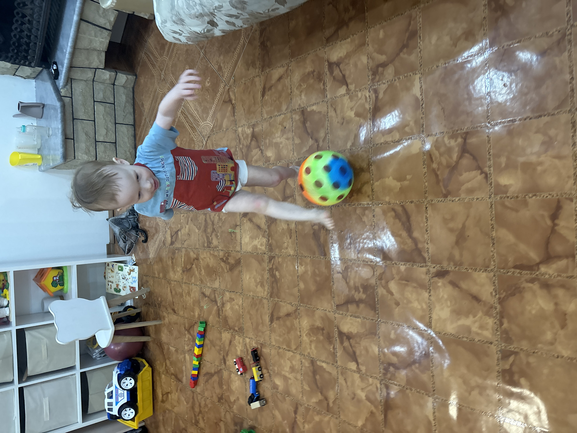 A child is playing with a ball in a room with tile flooring. The child is wearing a blue shirt and red overalls. The ball is multicolored. A shelf with toys and other items is visible in the background. Other toys are also located on the floor. The location appears to be a home interior. Location in Ferma, Russia cannot be determined.