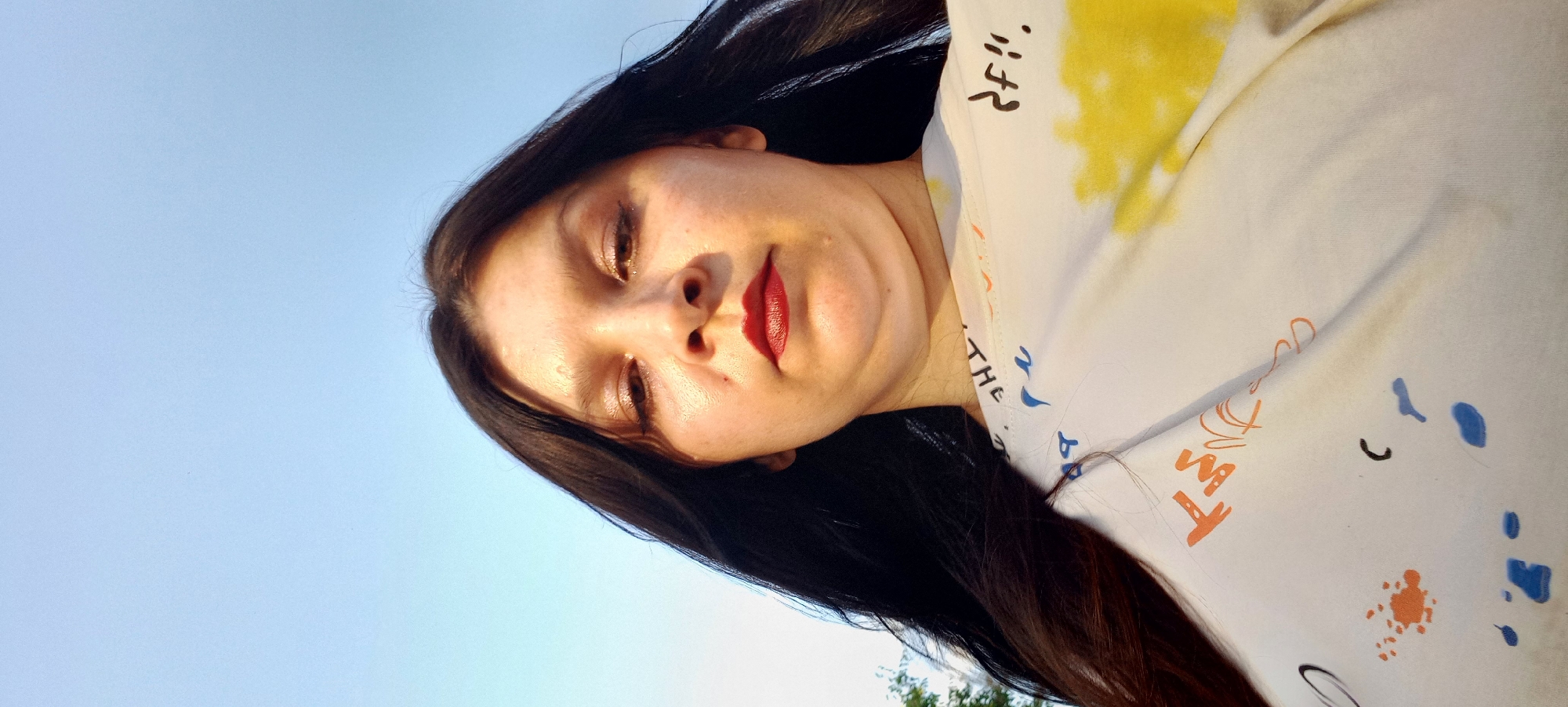 A low-angle, close-up, outdoor selfie shows a woman with long, dark hair and dark eyes. She is wearing a white t-shirt with colorful ink splatters and text printed on it. Her lips are painted with dark red lipstick. The background is a clear, pale blue sky. There is no information in the image to suggest a location in Klyntsi, Ukraine.