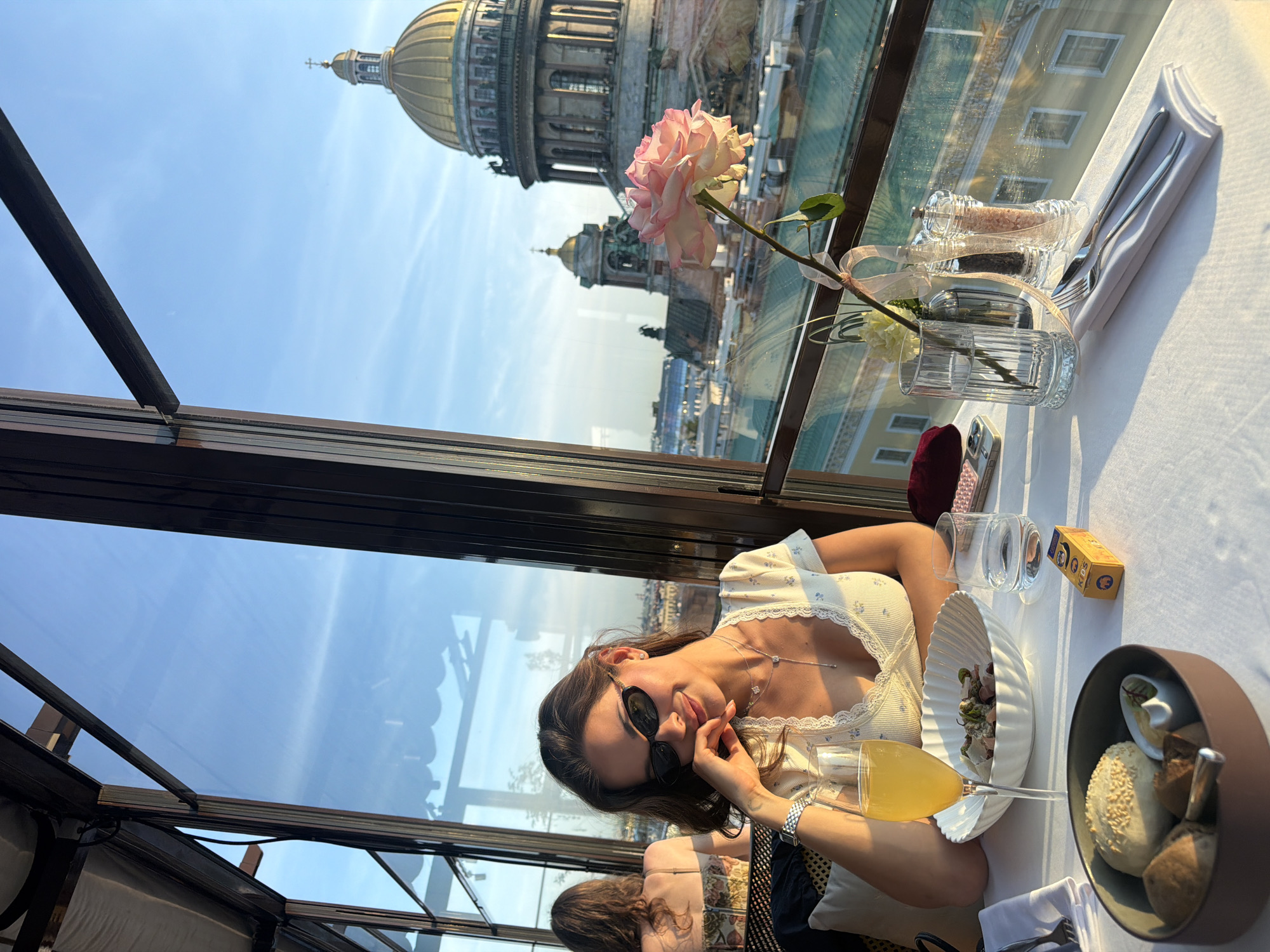 In the foreground, a woman with dark hair, wearing sunglasses and a white top with blue floral print, is seated at a table covered with a white tablecloth. She is smiling and resting her chin on her hand. In front of her is a glass of orange juice, a bowl of food, and a bread basket containing various rolls and butter. To her right are a glass of water, salt and pepper shakers, and a small yellow box. A single pink rose in a vase is positioned to the right of the frame, partially obscuring the view of the city.
The background reveals a cityscape viewed through a large window. Prominently visible is a large building with a golden dome topped with a cross, consistent with St. Isaac's Cathedral in St. Petersburg, Russia, not Lednevo. Other buildings and the sky are also visible. The lighting suggests it is late afternoon or early evening. Another person with brown hair is partially visible in the upper left corner of the image, seated further back at another table.