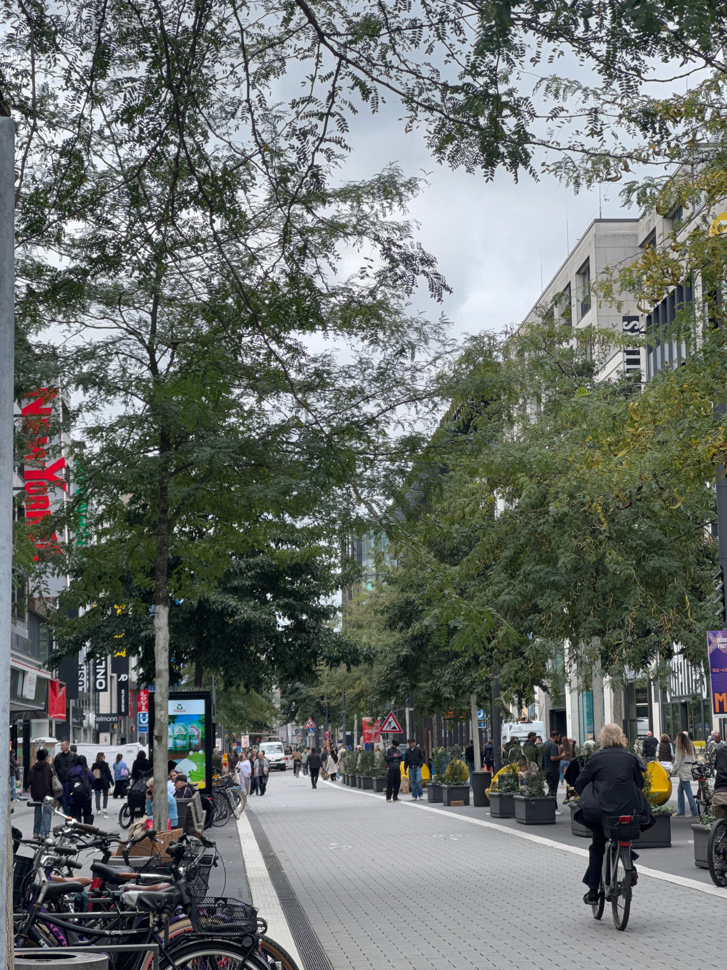 The image depicts a street scene in Dusseldorf, Germany, likely during daylight hours under an overcast sky. The street is paved with gray interlocking bricks and lined with trees and planters. On the left side of the street, buildings house various shops with prominent signage, including "ONLY" and what appears to be "NEW YORK". A large digital screen displays a frog character. On the right side of the street, modern buildings are visible.
The street is populated with a number of pedestrians and cyclists. Several people are walking along the sidewalk, while others are seated at an outdoor cafe area. Bicycles are parked in the foreground on the left, and a woman is riding a bicycle in the middle ground on the right. The overall atmosphere suggests a busy urban environment with a mix of commercial activity and pedestrian traffic.
