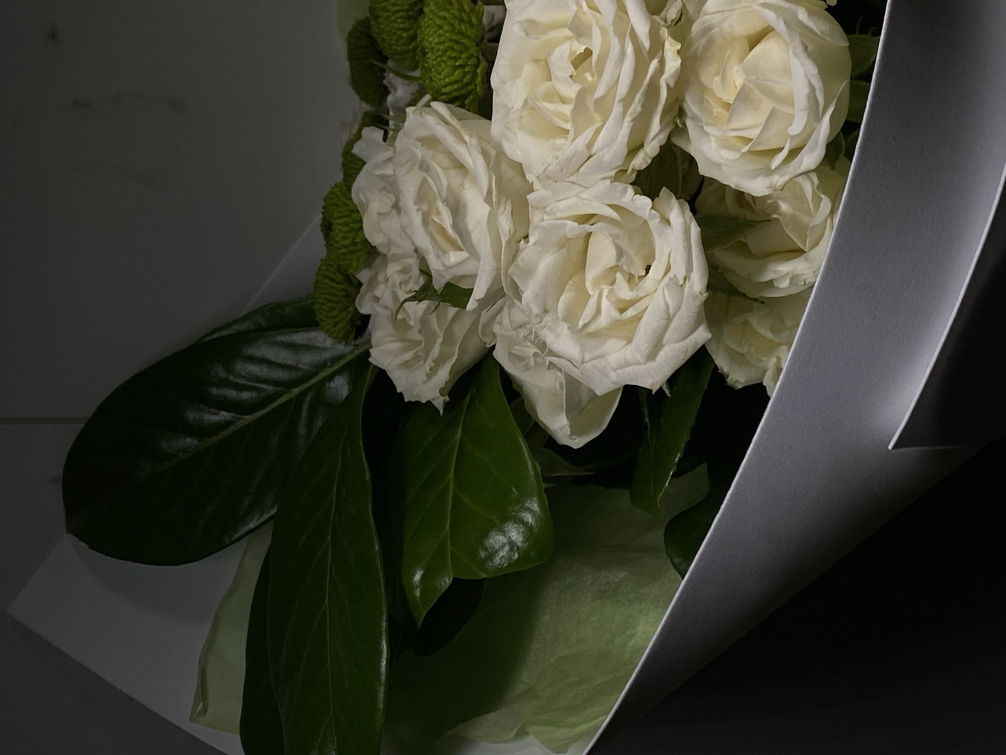 A bouquet of white roses and green chrysanthemums is presented in a minimalist white and grey paper wrapping. The flowers are arranged with large, dark green leaves, creating a dramatic contrast against the pale blooms. The wrapping paper is folded in a way that creates sharp, geometric lines and shadows, adding to the contemporary aesthetic. The background is dimly lit, with a hint of a marble texture on the right side, suggesting an indoor setting that is elegantly appointed. There are no people visible in the image. The overall impression is one of sophisticated simplicity. The image does not contain any information that would allow for determining a specific location in the city of Starokorsunskaya, Russia.
