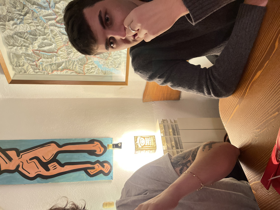 The media file presents an indoor, candid photograph rotated 90 degrees counter-clockwise. The primary subject is a young adult male, positioned on the top and left side of the frame, looking directly at the camera with a neutral expression. He has dark hair and dark eyes, and is wearing a dark grey knitted sweater. His right hand is near his face, with a simple silver ring visible on his ring finger.
A second person is partially visible on the bottom and right side of the frame. Their right arm and shoulder are in view, revealing a dark, intricate tattoo, possibly of a dragon or similar creature, on their upper arm. They are wearing a light grey t-shirt and a gold-colored chain bracelet on their wrist. Dark, curly hair is also visible at the very bottom edge.
Both individuals appear to be seated at a sturdy wooden table with a visible grain, which occupies a significant portion of the lower and right side of the image. On the table, near the second person's arm, is a small, red item, possibly a gift box.
The background wall features several decorative elements. Above the male subject's head (when correctly oriented), a framed topographic map depicting mountainous terrain with blue and red lines hangs. Below this, a long, rectangular, light blue painting with a stylized, peach-colored human figure is mounted. Two bottle necks, seemingly from liquor bottles, are integrated into the wall as light fixtures, with one appearing to pierce the painting. A square wall light bearing the text "Jack Daniel's" is prominently visible further right, emitting a strong glow. Adjacent to this light, a white, multi-compartment shelving unit is partially seen. A small mark, "V°", is visible on the lower right of the blue painting.
The lighting is artificial, suggesting the photo was taken in the evening or at night. The overall atmosphere is casual and relaxed, likely depicting a social gathering in Sondrio, Italy.