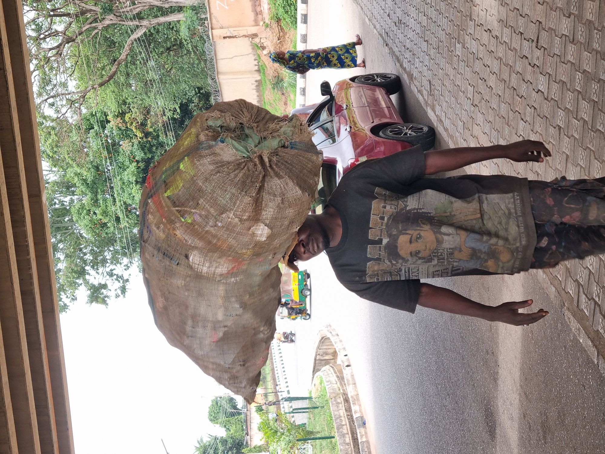 A man in Jos, Nigeria, carries a large burlap sack on his head while walking near a street under an overpass. The man wears a dark t-shirt with a print and patterned pants. His hands are outstretched. In the background, a red car and a person in colorful clothing are visible. A yellow and green motorized tricycle is also present in the distance. The setting includes trees, buildings with barbed wire, and paved surfaces with patterned blocks. The sky is bright.