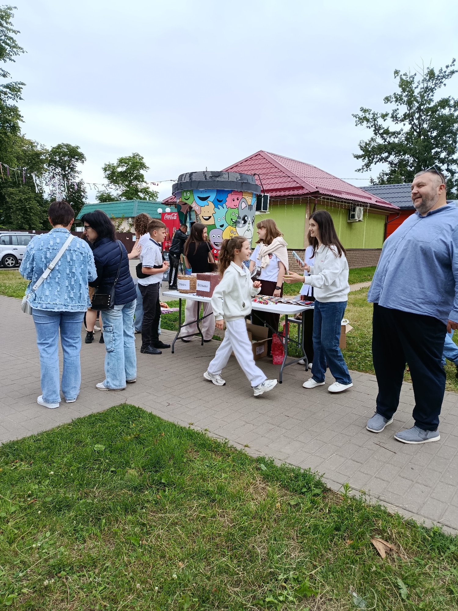 An outdoor scene in Smilyavitski Sel'ski Sovet, Belarus, features a gathering of people around a table laden with goods. A diverse group, including adults and children, are engaged in various activities. Some individuals are standing, observing, or interacting with items on display. A young girl in a white tracksuit is in motion, walking past the table. Another woman, dressed in a white hoodie and jeans, is holding a phone, possibly in the process of making a purchase or sharing information. A large man in a blue shirt and dark pants stands prominently in the foreground, looking to his right and smiling. Behind the table, several other individuals, including a young boy in a white shirt and dark pants, are present. The background reveals a building with a red roof, a vibrant, cartoon-decorated structure resembling a large trash can, and trees. The ground is a paved walkway bordered by grass. The overall atmosphere suggests a casual outdoor event or market.