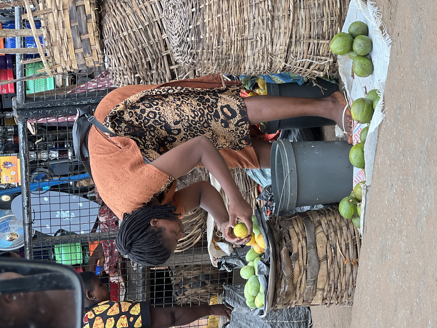 This media file captures a vibrant scene at an outdoor market in Nkpor, Nigeria.
The primary subject is a woman standing, slightly bent over, actively tending to her produce stall. She is wearing an orange top and a black and brown leopard-print skirt or dress. Her hair is styled in dark dreadlocks or braids, and she is barefoot. A black shoulder bag is slung over her left shoulder. She holds a yellow fruit in her hands, appearing to sort or arrange the items within a large, round woven basket positioned on the ground in front of her.
Her stall features a variety of green and yellow fruits, likely mangoes or similar, displayed prominently. These fruits are arranged in several large, rustic woven baskets and also spread out on a white sackcloth laid directly on the ground. Two dark grey plastic buckets are visible as part of her setup, one behind her and another near the fruit display. A single white flip-flop lies near her right foot.
The background reveals a bustling market environment. Intricately woven bamboo or wood structures form parts of other stalls or storage units. Various other goods, including what appear to be fans and other electronic items, are stacked on shelves or stands in the deeper background. The word "Commox" is discernible on a product box in this area. A partial view of another individual, possibly a child or another vendor, wearing a yellow and black patterned top, is visible in the lower left portion of the image, partially obscured by a reflection. The overall scene suggests a typical daytime market setting, characterized by active commerce and local produce.