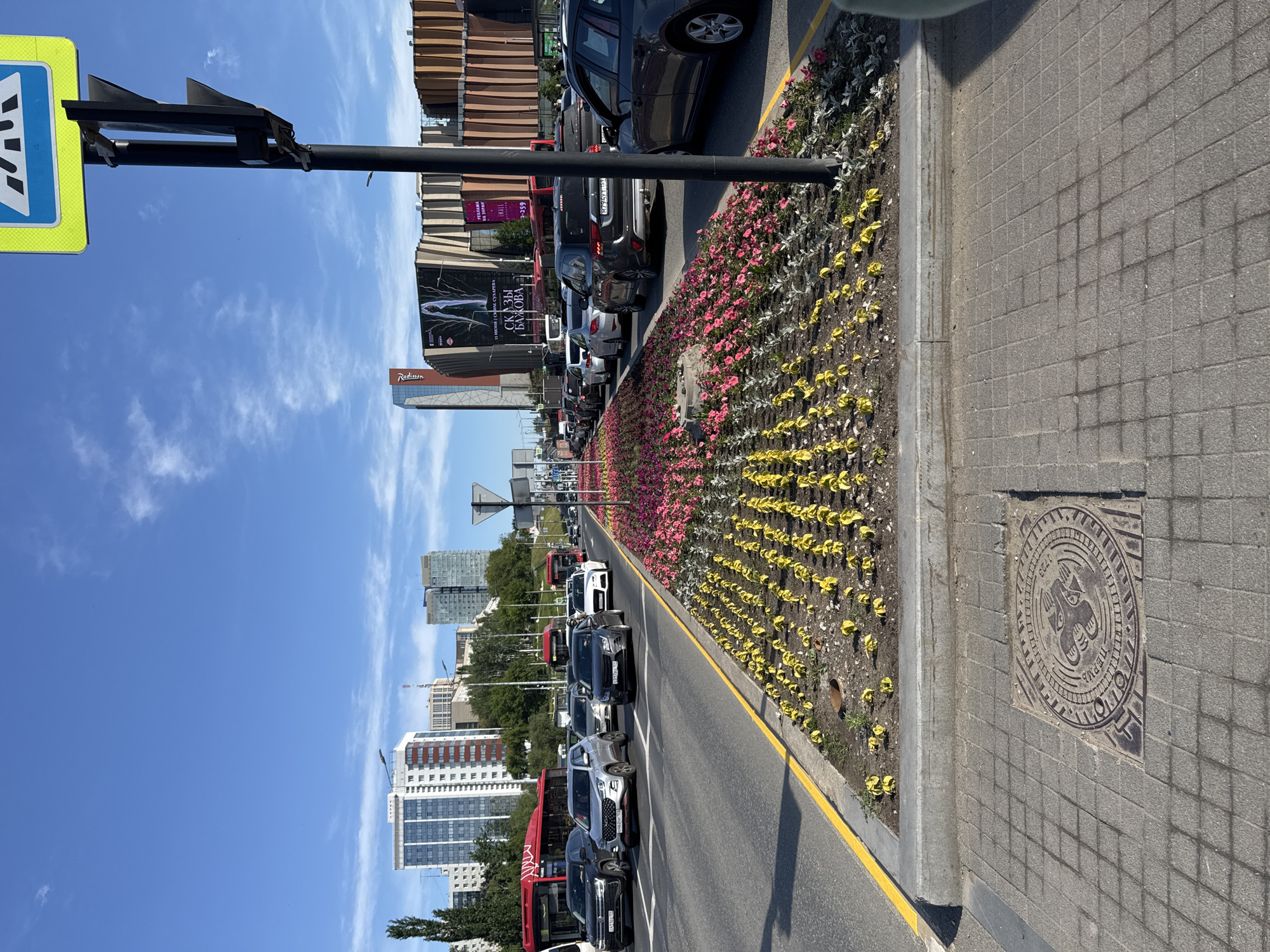 The image displays a bustling urban street scene in Perm, Russia, under a clear blue sky with sparse white clouds, indicative of a bright, sunny day.
In the foreground, a grey tiled sidewalk runs parallel to a vibrant flower bed. Embedded in the sidewalk is a circular manhole cover, intricately designed with what appears to be the Perm Bear, a symbol from the city's coat of arms, along with the text "1723" and "ПЕРМЬ" (Perm). The adjacent flower bed features rows of vivid pink and red blossoms closer to the street, transitioning to rows of young, bright yellow-green plants further back.
The flower bed borders a multi-lane asphalt road, edged with a solid yellow line. A line of vehicles, including various cars such as SUVs and sedans, and distinctive red city buses, suggests either heavy traffic or parked cars.
To the left, a black street pole holds a blue and yellow pedestrian crossing sign. Beyond the road, several prominent buildings define the cityscape. A large building with a textured brown facade displays a billboard featuring Russian text: "11 ИЮНЯ В ОПЕРЕ СКАЗЫ БАЖОВА" (11 June in the Opera Bazhov's Tales). A smaller sign above it reads "РЕКЛАМА НА ЭКРАНЕ 2240 3359" (Advertising on the screen 2240 3359). Nearby, another building clearly shows the "Radisson" brand. In the distant background, a cluster of modern high-rise buildings with glass facades completes the urban panorama. The scene captures a typical, active city environment, emphasizing well-maintained public spaces and a clear view of architectural elements.
