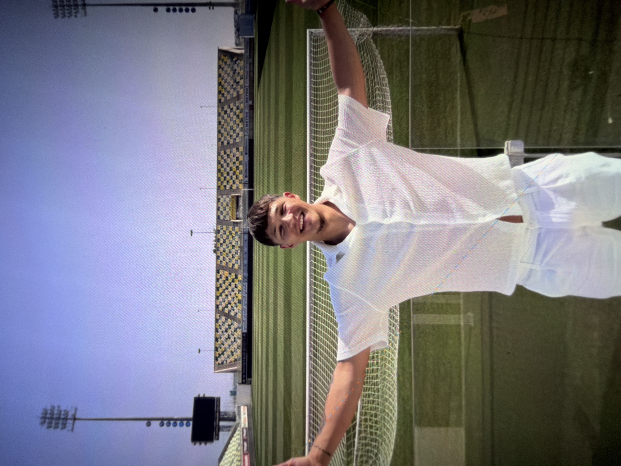 A young man stands in front of a soccer stadium, smiling and with his arms outstretched. He is wearing a white short-sleeved collared shirt and white pants. The stadium has green grass, a soccer net, and yellow and black seats. There are also tall light poles and a large screen. The sky is a clear blue. The image appears to be taken on a sunny day.
