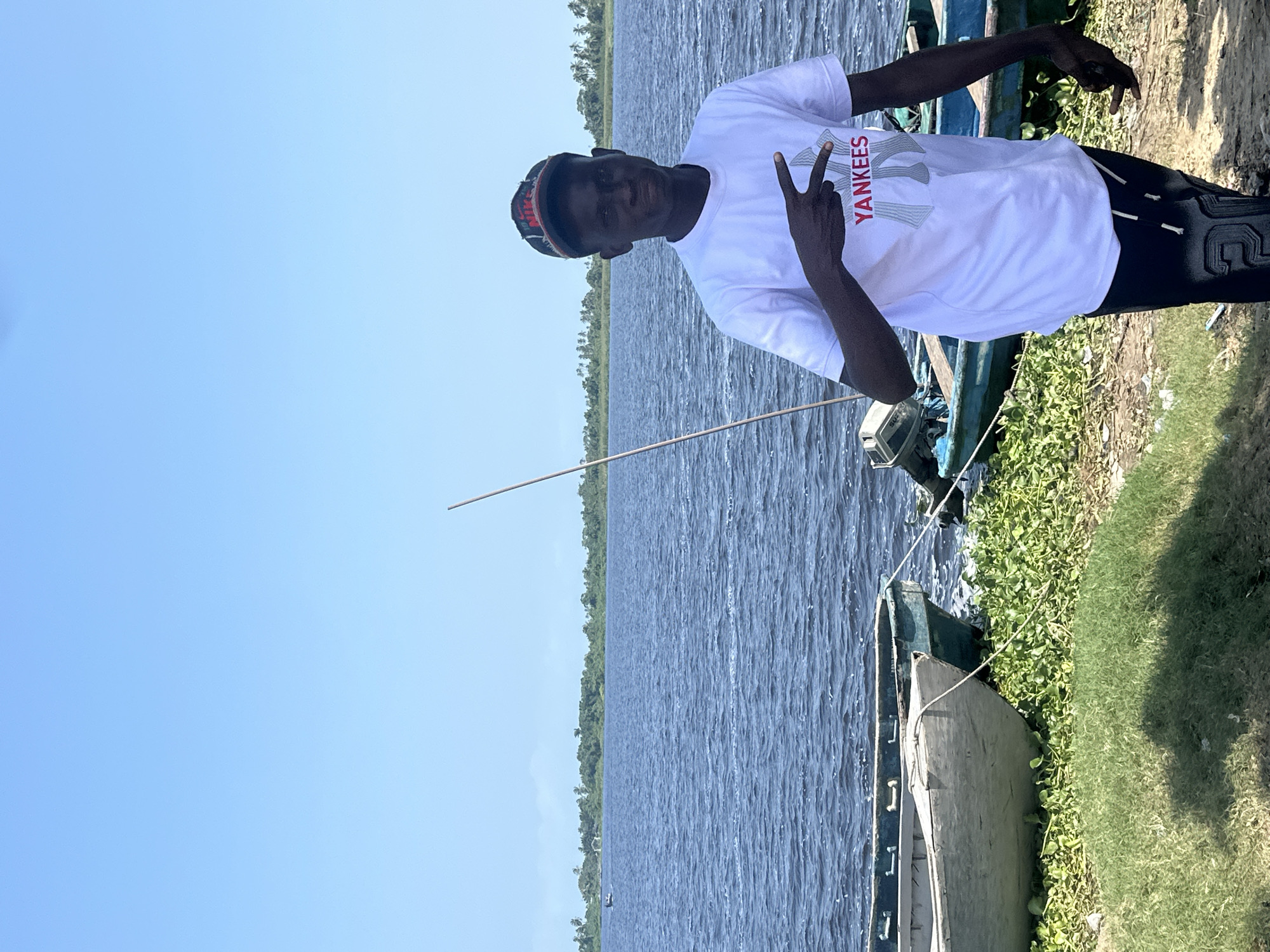 The image shows a young Black male standing on the bank of a body of water, likely a lagoon or river, in Badagry, Nigeria. He is wearing a white t-shirt with the word "YANKEES" written in red and grey letters, and a patterned cap. The man is looking towards the camera and holding up two fingers in a peace sign. He appears to be smiling.
Behind the man, several boats are docked along the shore. The water is calm, with small ripples on its surface, and a lush green landscape with palm trees lines the opposite bank. The sky is a clear, bright blue, suggesting it is daytime with good weather. The overall impression is one of a relaxed, outdoor scene. The presence of boats and the natural landscape point to a location near the water, common in coastal areas like Badagry.