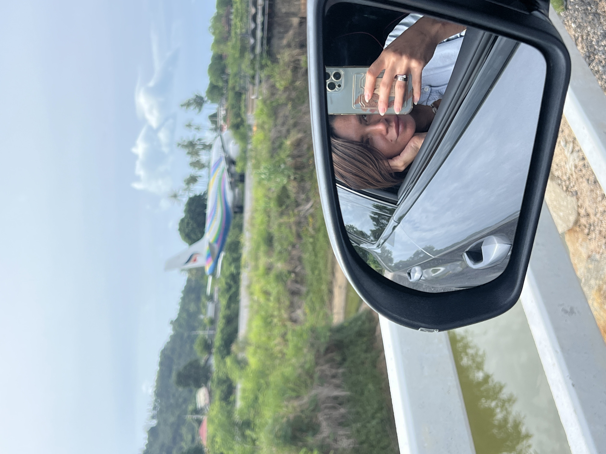 An over-the-shoulder medium shot shows a woman taking a selfie in the side mirror of a vehicle. She is leaning her head on her hand, with a slight smile. The woman is wearing a light blue button-down shirt and a ring on her left hand. A colorful airplane is visible in the background, on a runway, with lush green trees and hills surrounding it. The sky is overcast with a few clouds. The vehicle's interior is reflected in the mirror, along with the woman.