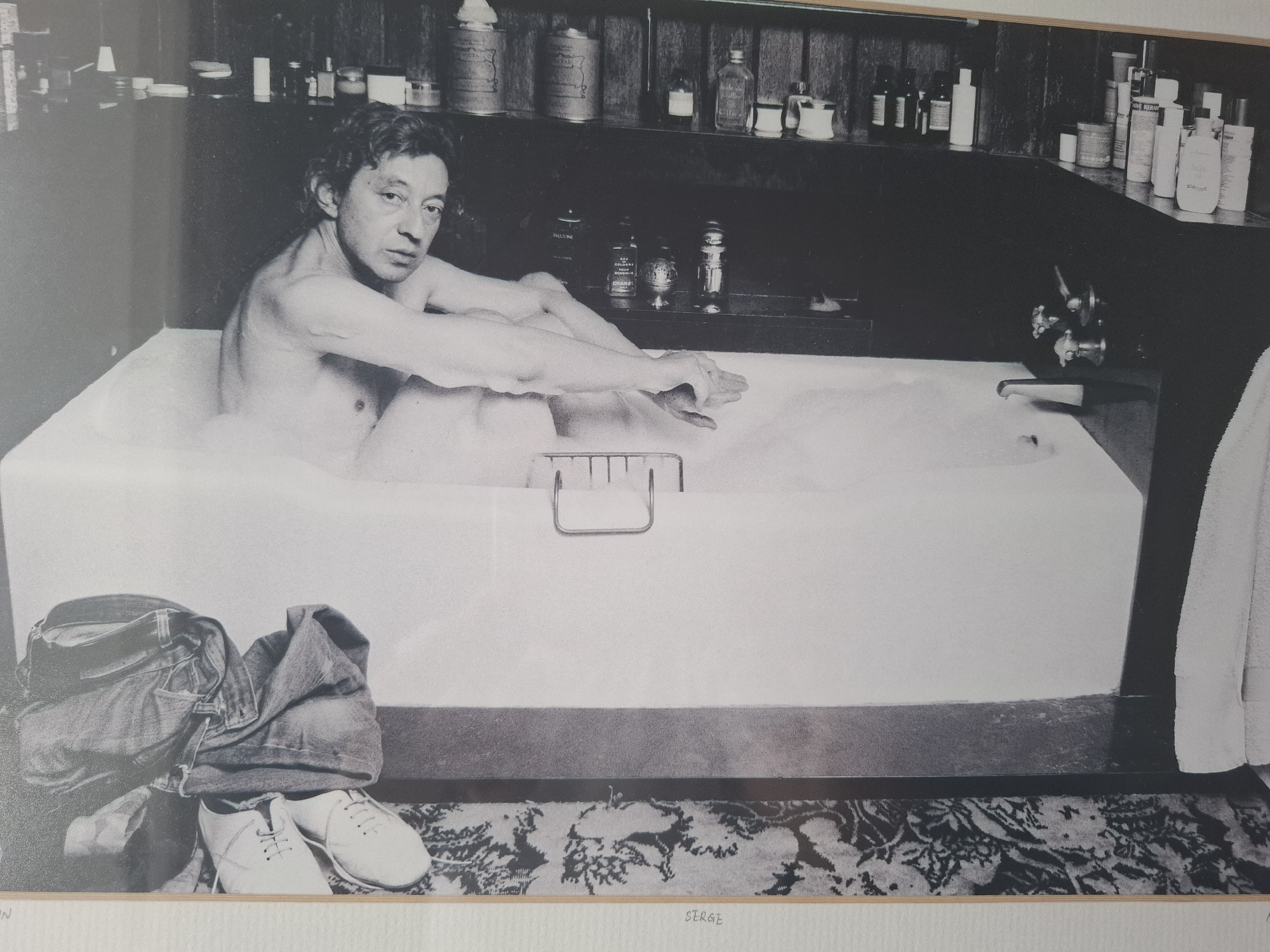 The black and white photograph shows Serge Gainsbourg in a bathtub filled with bubbles. He is partially submerged, with his arms resting on the edge of the tub. His gaze is directed towards the camera. The background features shelves lined with various bottles and containers, suggesting a bathroom setting. To the left of the bathtub, a pair of jeans and white sneakers are placed on a patterned rug. The image is a close-up of Gainsbourg and the tub, with the shelves and clothing visible in the periphery. The location is not discernible from the image itself, and therefore, any mention of Montreuil, France is not supported by the visual information provided.