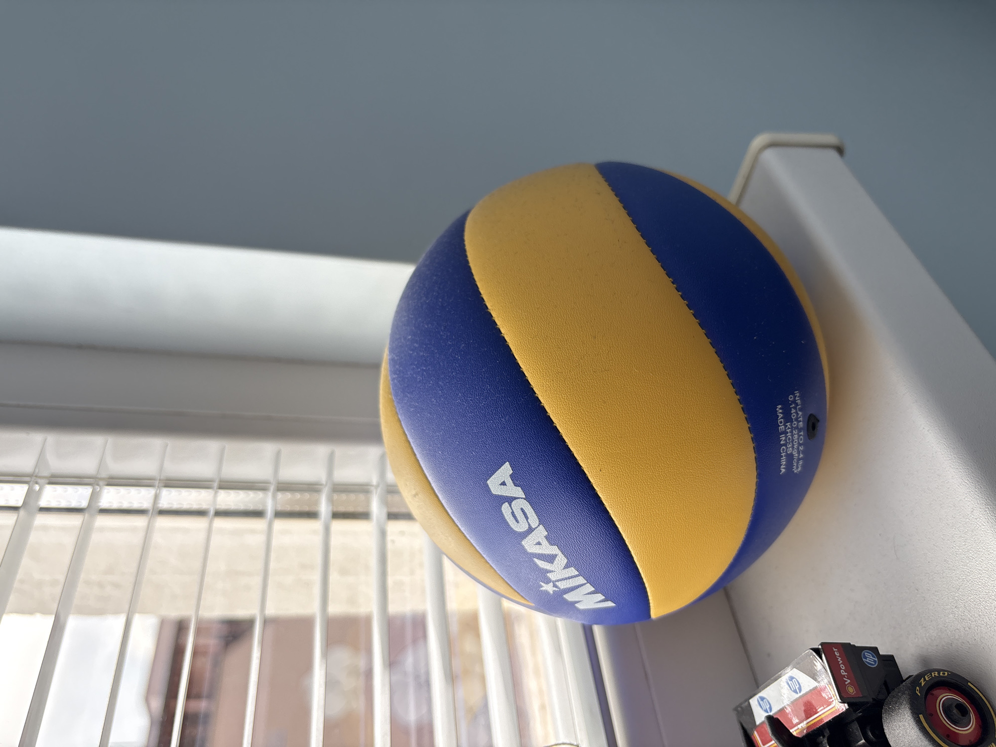 An indoor, eye-level shot captures a blue and yellow Mikasa volleyball resting near a window with white venetian blinds, possibly on a windowsill or shelf. The volleyball dominates the right side of the frame, angled towards the viewer, showcasing its textured surface and vibrant colors. The "MIKASA" logo is prominent in white lettering on a blue panel. Closer inspection reveals manufacturing details printed on the ball: "INFLATE TO 2-4 LBS", "0.140-0.280kgf/cm²", "KHC3S", and "MADE IN CHINA".
To the bottom right, partially obscured by the volleyball, is a small toy car or truck. Its black body features red and white accents, with visible "V-Power" and "HP" logos. One of its wheels, bearing the "P ZERO" brand, is distinctly visible.
In the background, behind the volleyball and above the blinds, a light blue-gray wall or ceiling is visible. Bright natural light streams in from the window, highlighting the subjects and casting subtle shadows. The blinds are slightly open, revealing a blurry, light-colored background outside, possibly buildings or an abstract outdoor scene. The overall impression is a casual, well-lit still life. The scene is static, with no discernible activity taking place. The time of day appears to be daytime due to the bright, natural light. The provided context for the location is Sofia, Bulgaria.